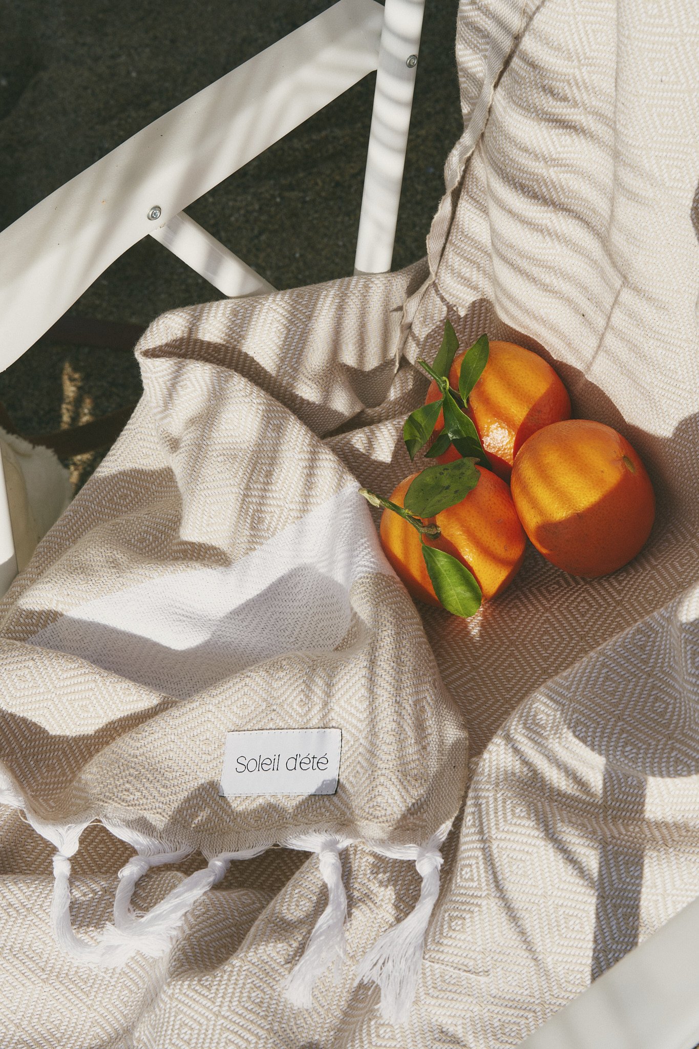Three oranges with green leaves resting on a beige patterned fabric labeled "Soleil d'été," placed on a white chair.