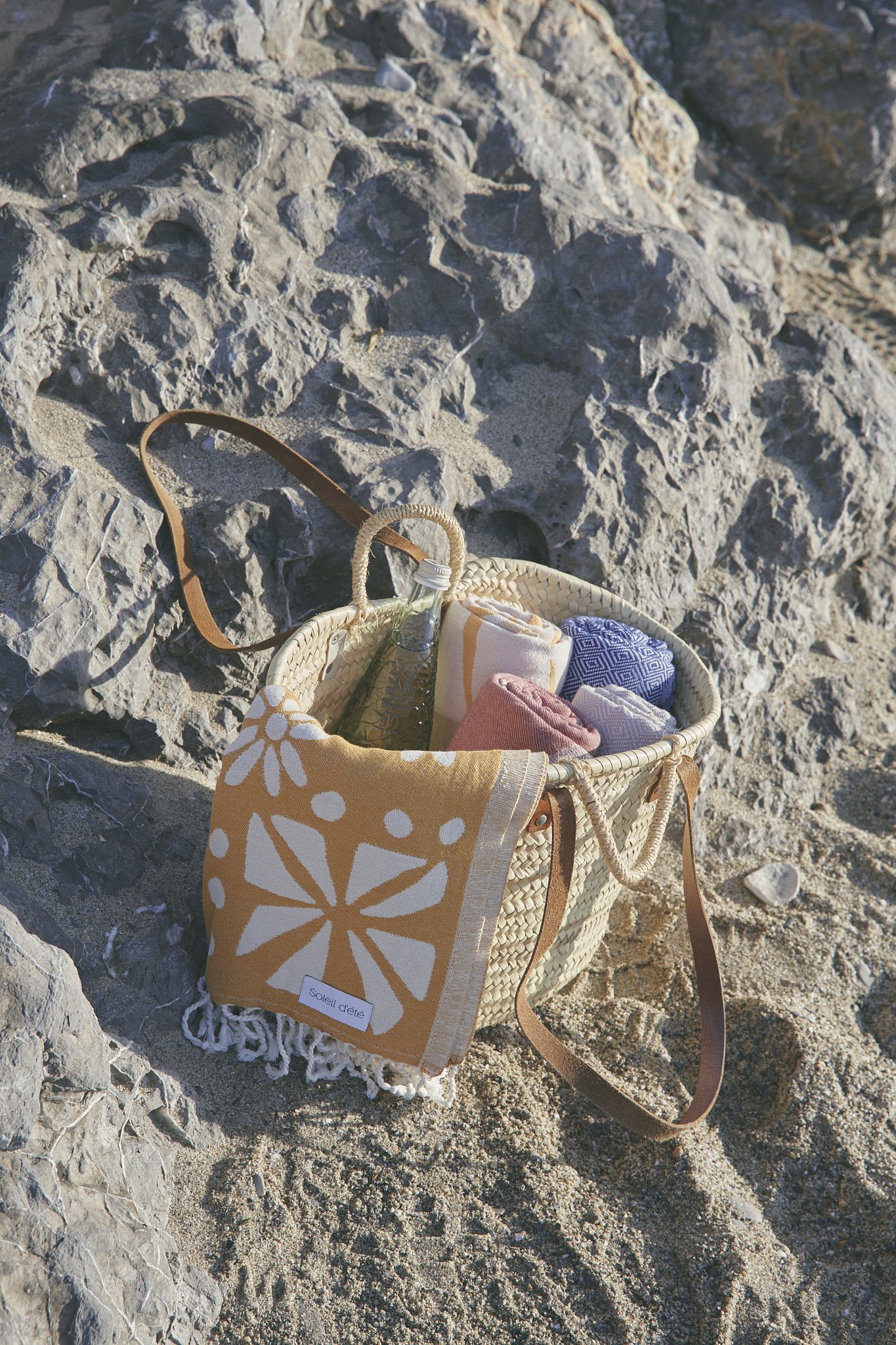 Beach scene with a woven bag containing a bottle, towel, and cloth on rocks.