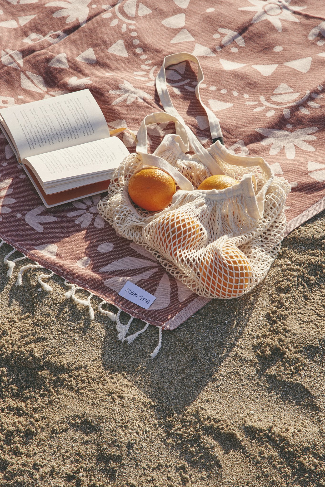Beach scene with a patterned towel, open book, mesh bag containing oranges, and sand background.