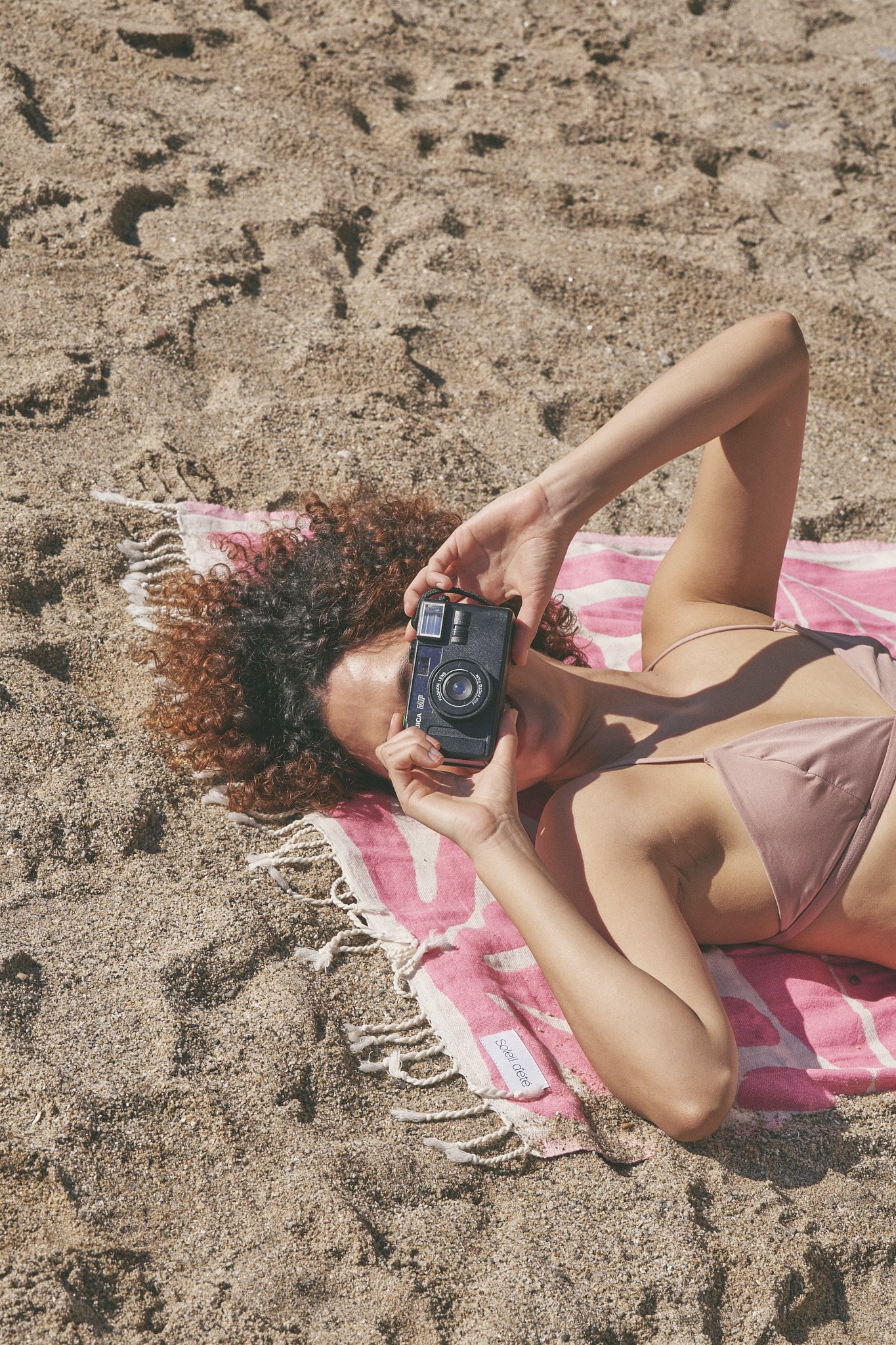 Person lying on beach with a camera, resting on a pink towel.