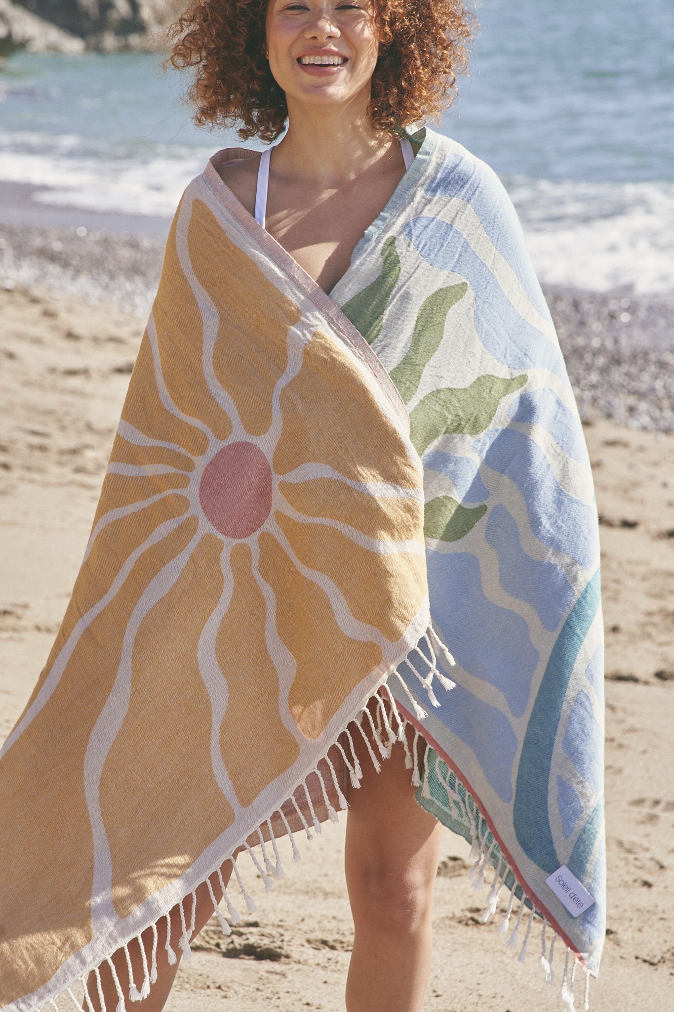 Person at the beach wrapped in a colorful towel with sun and wave patterns.