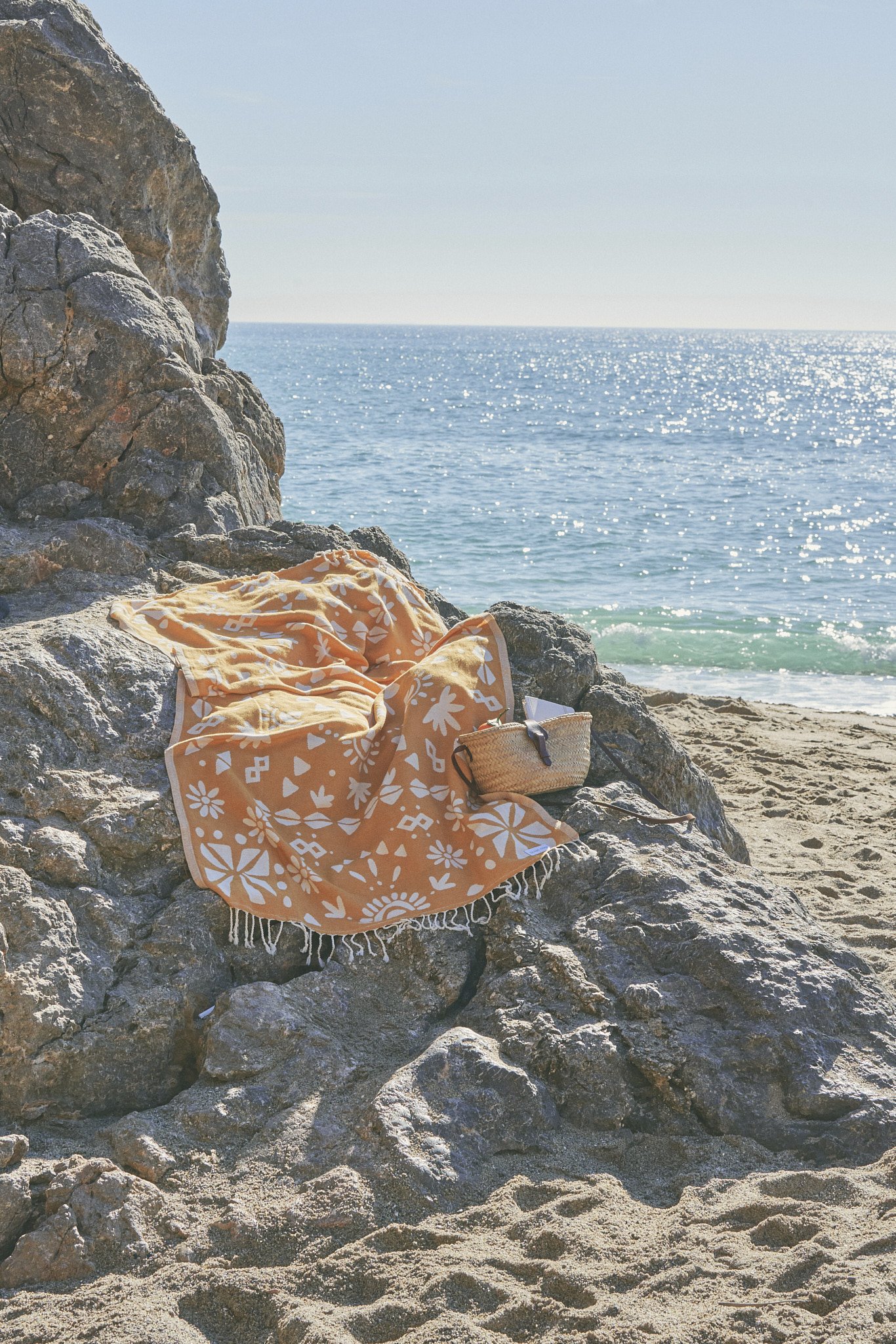 A patterned beach towel and a woven basket on a rocky area by the ocean with a sandy beach.