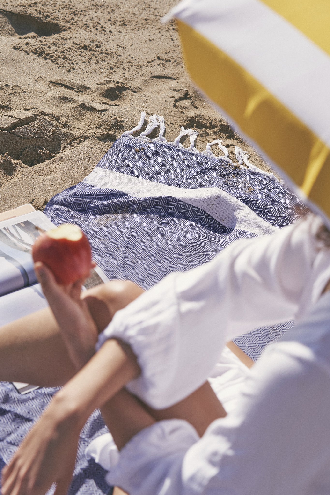 Person relaxing at the beach, sitting on a blue patterned towel, holding an apple, with part of a yellow beach umbrella visible.
