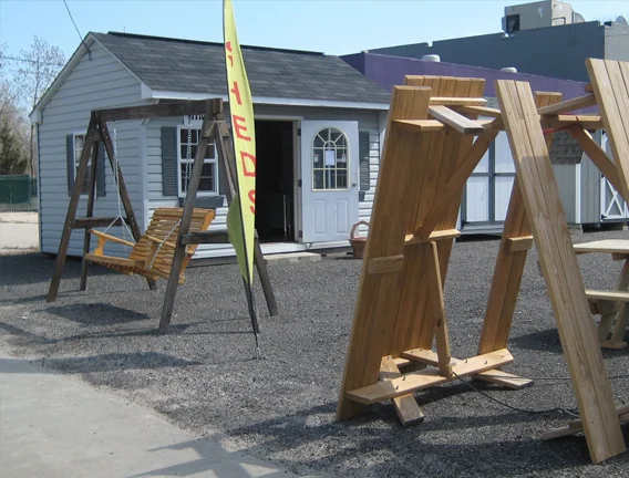 Outdoor scene with a small white building, wooden outdoor furniture, and construction wood pieces. A yellow flag with red text is visible.