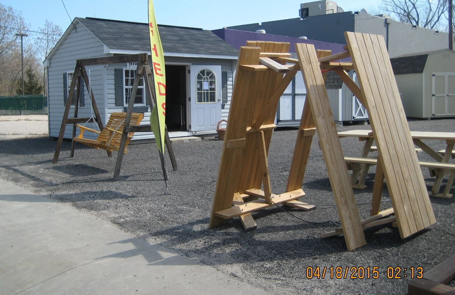 Outdoor scene with wooden swings, picnic tables, and storage sheds in a gravel lot, with clear sky and trees in the background.