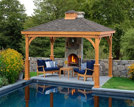 Outdoor gazebo with a fireplace, surrounded by garden plants, overlooking a swimming pool.