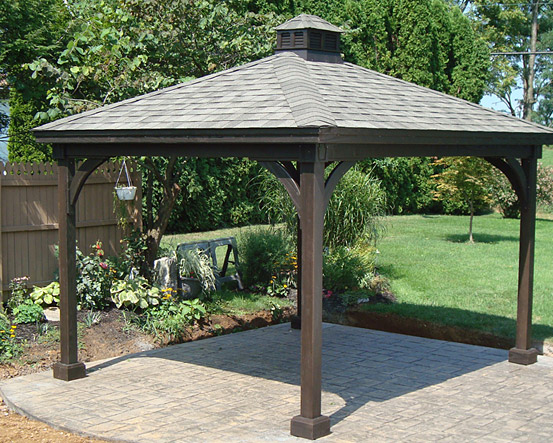 A wooden gazebo with a shingled roof in a backyard, with a sitting area and plants underneath.