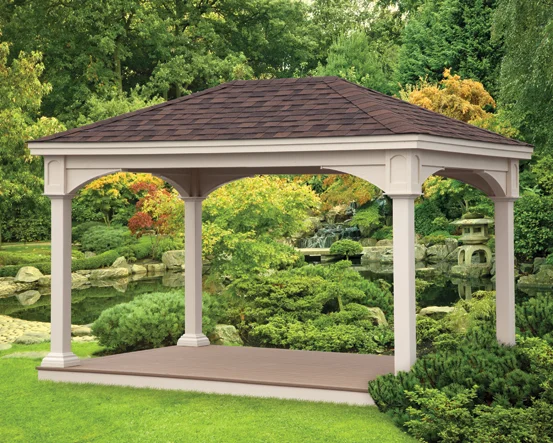 A white gazebo with a brown roof in a lush garden with trees, bushes, rocks, and water features.