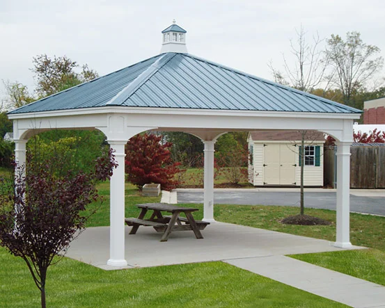 A white gazebo with a blue metal roof and a cupola, situated on a concrete pad in a park-like setting with grass, trees, and a picnic table inside.