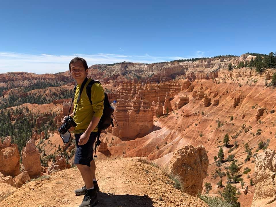 A person standing on a mountain trail at Bryce Canyon, with red rock formations and evergreen trees in the background under a clear blue sky.