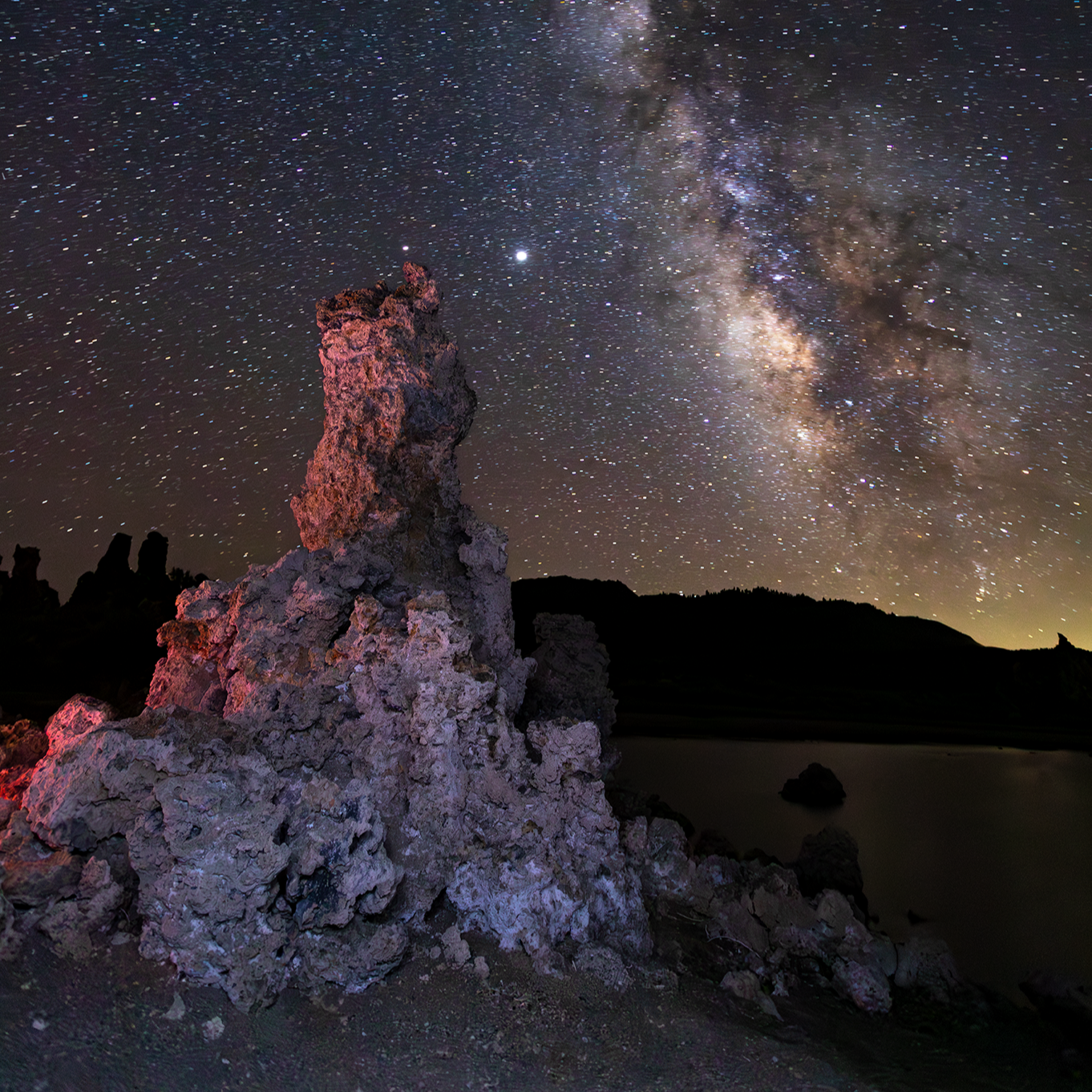 Night sky filled with stars and the Milky Way galaxy above rocky formations and a body of water in the distance.