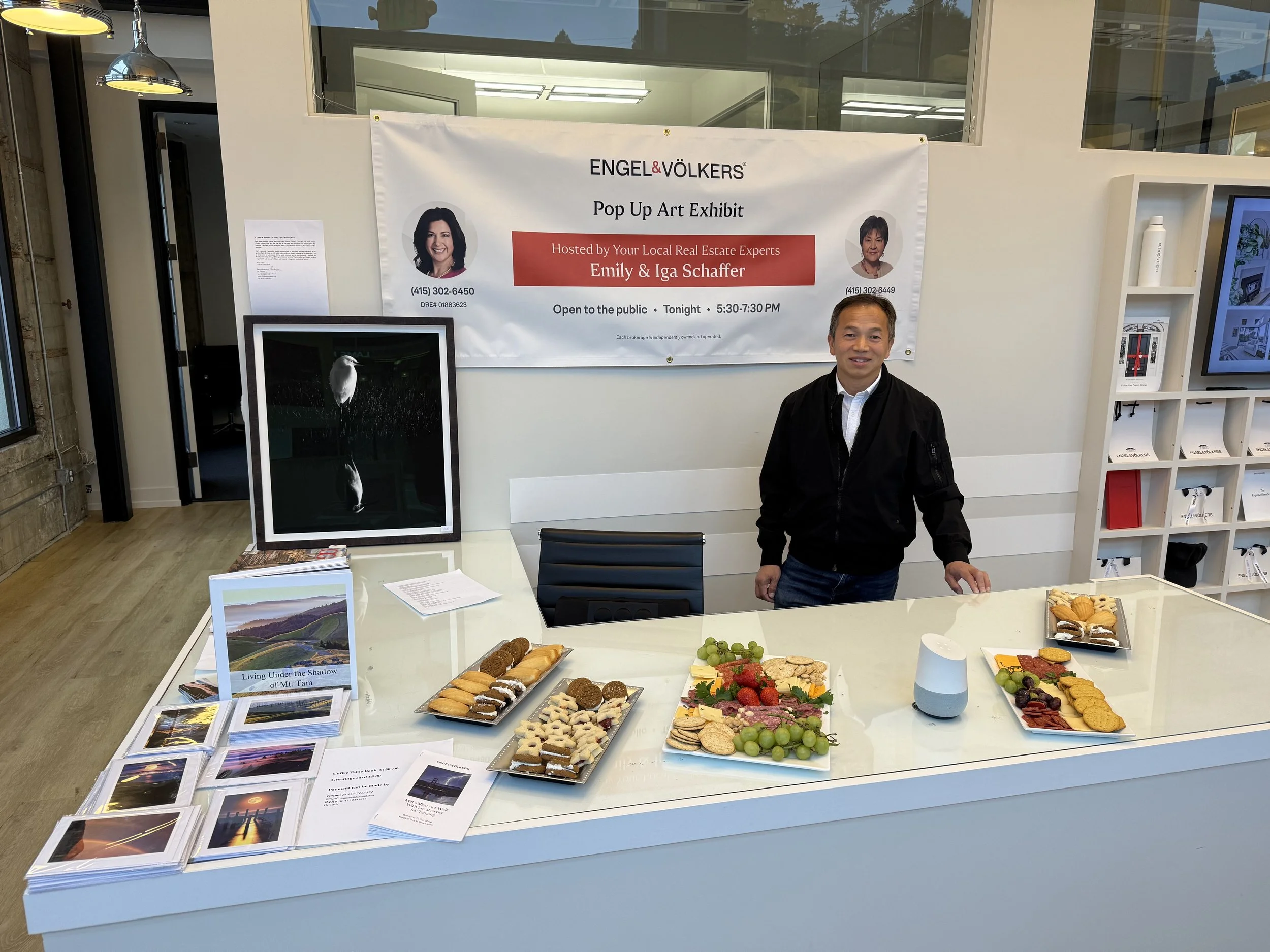 A man standing behind a white table with assorted snacks and brochures at an art exhibit event. A large banner behind him displays the names Emily and Iga Schaffer, with photos of two women, and the event details.