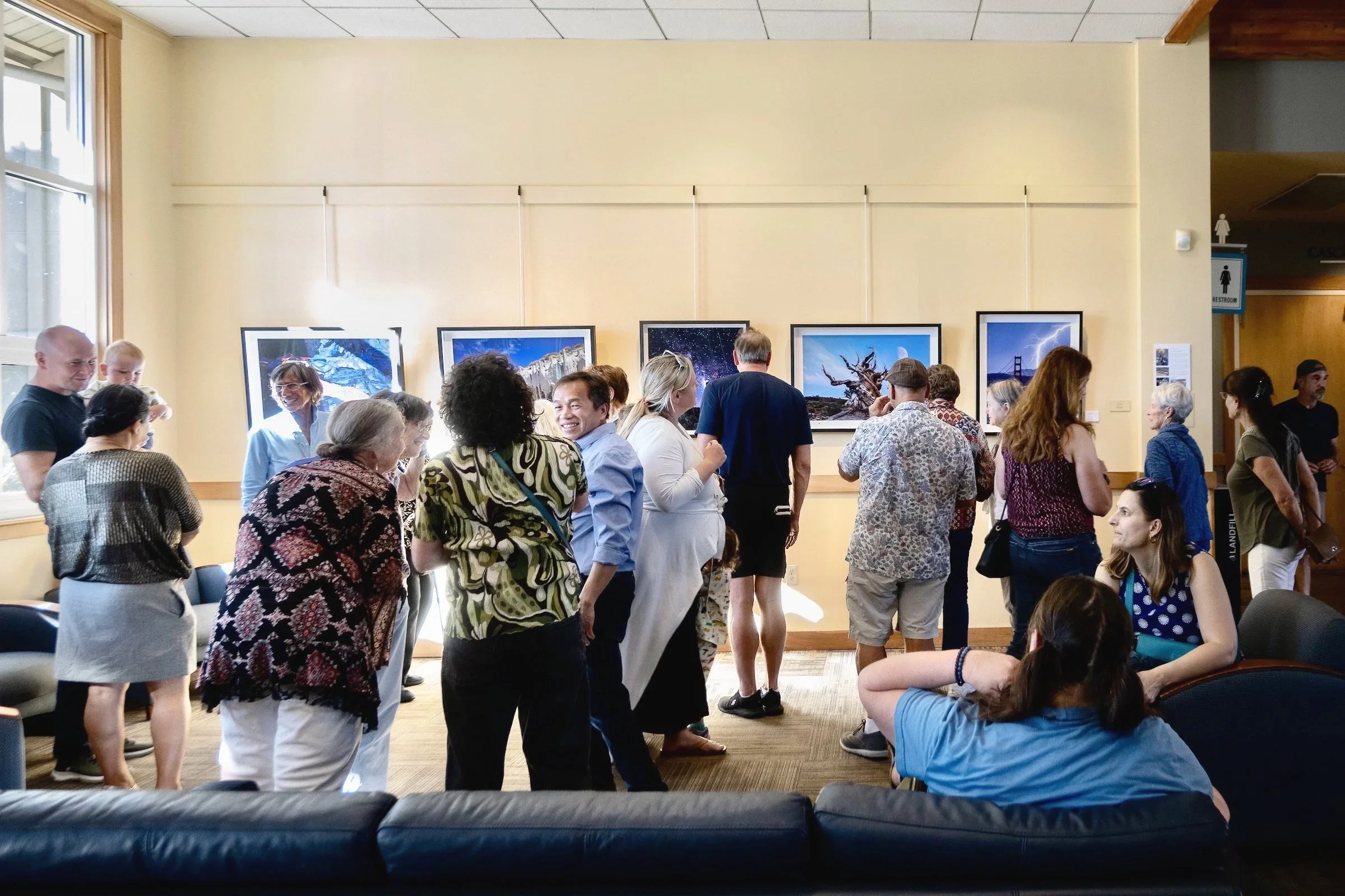 People gathered at an art gallery viewing photographs displayed on the wall. Some are looking at the pictures, others are conversing or sitting. Natural light from large windows illuminates the room.