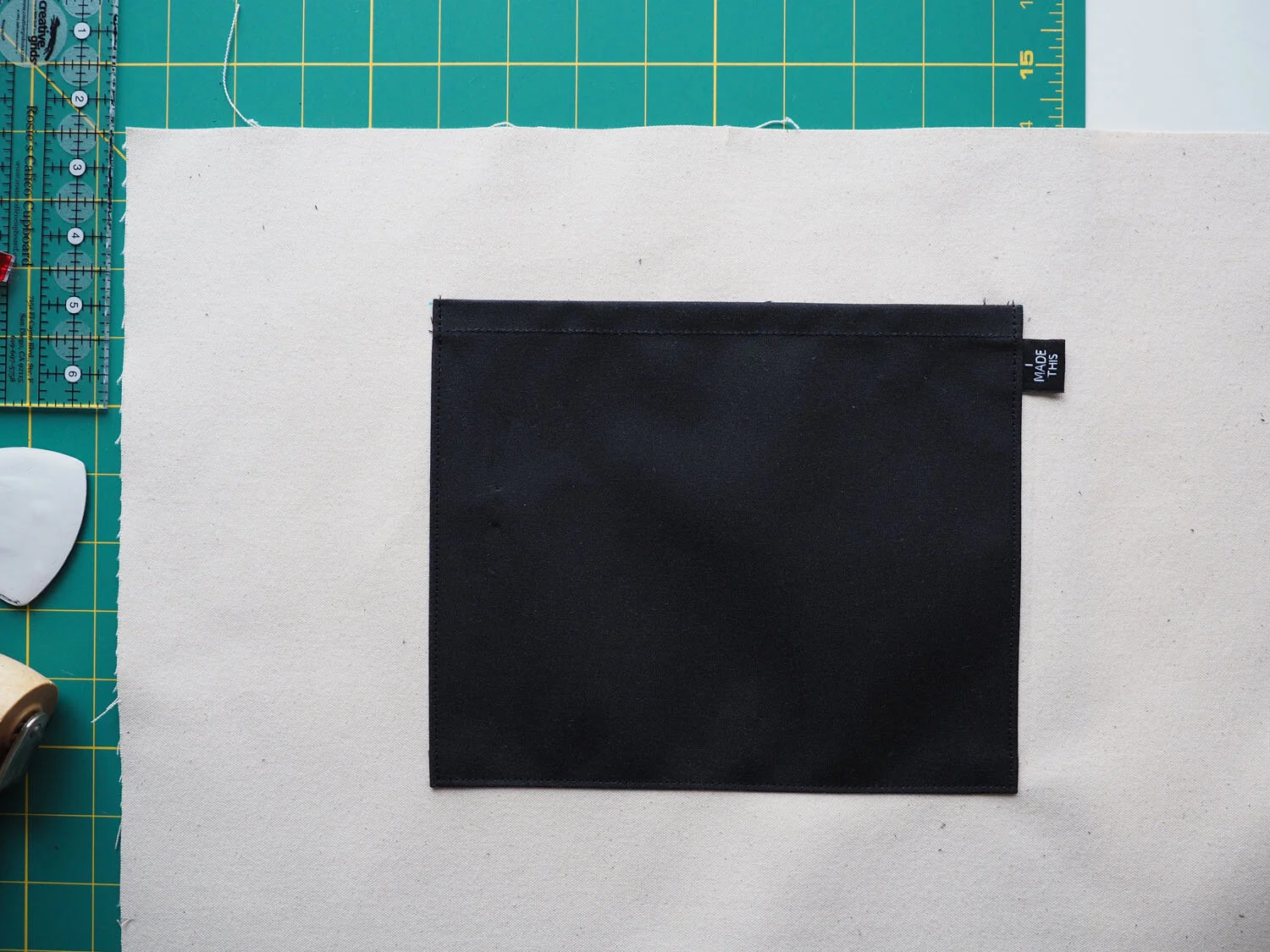 A black waxed canvas pocket sewn on a piece of natural canvas. An “I made this” label peeks out from one edge of the pocket. 