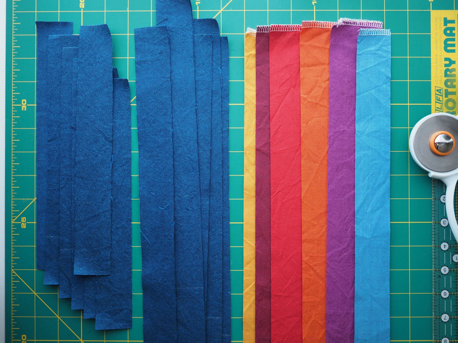  Precut strips of navy, red, yellow, orange, blue, and purple fabric laid flat on a cutting mat  