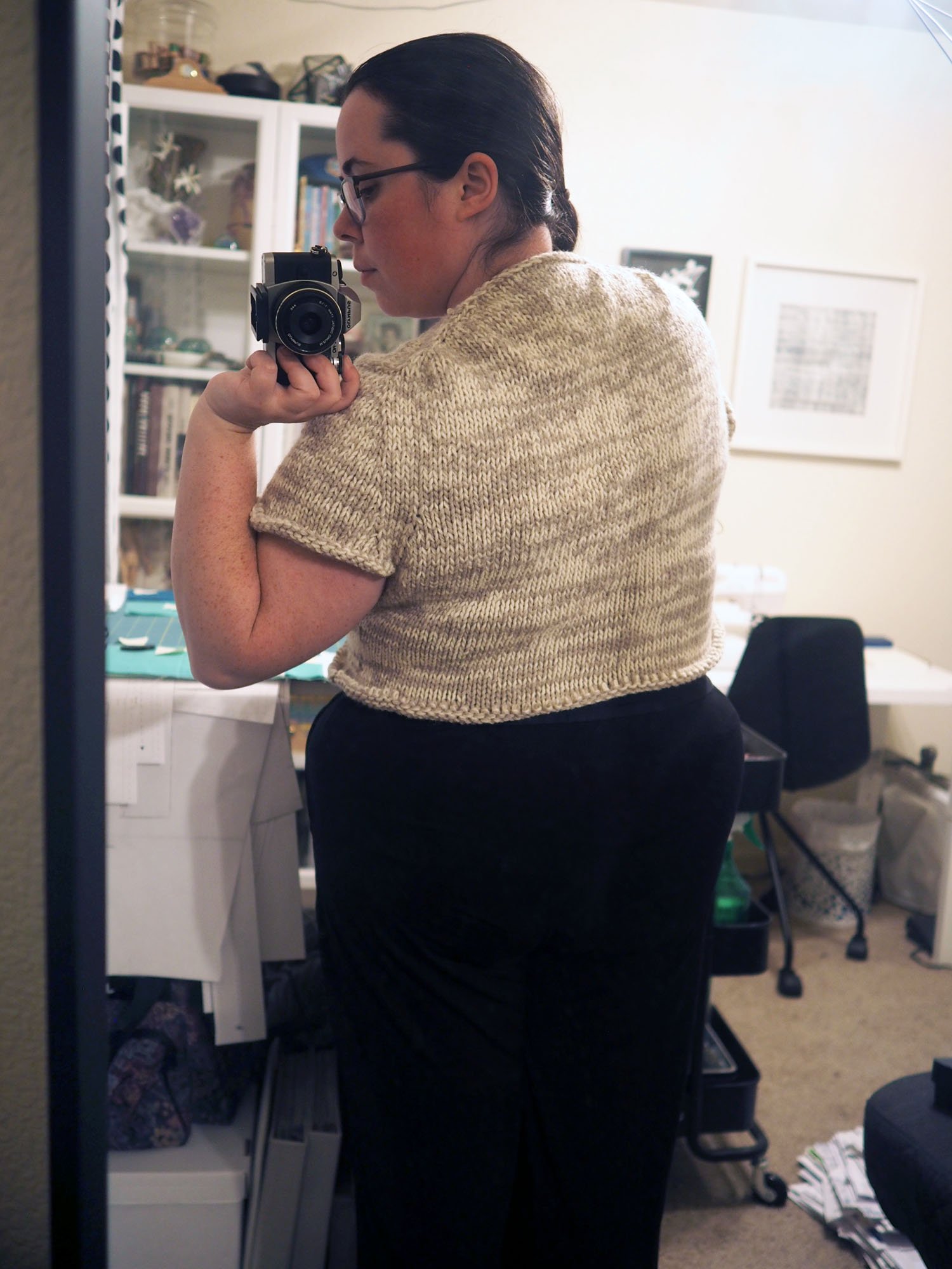 A white woman takes a selfie of her back in a mirror while wearing a partially finished grey and cream v-neck sweater
