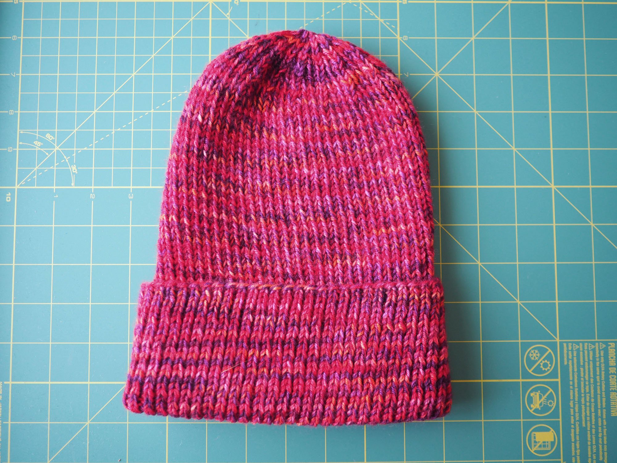 A knit hat with subtly variegated striping in pink, red, orange, and purple is laid flat on a cutting mat