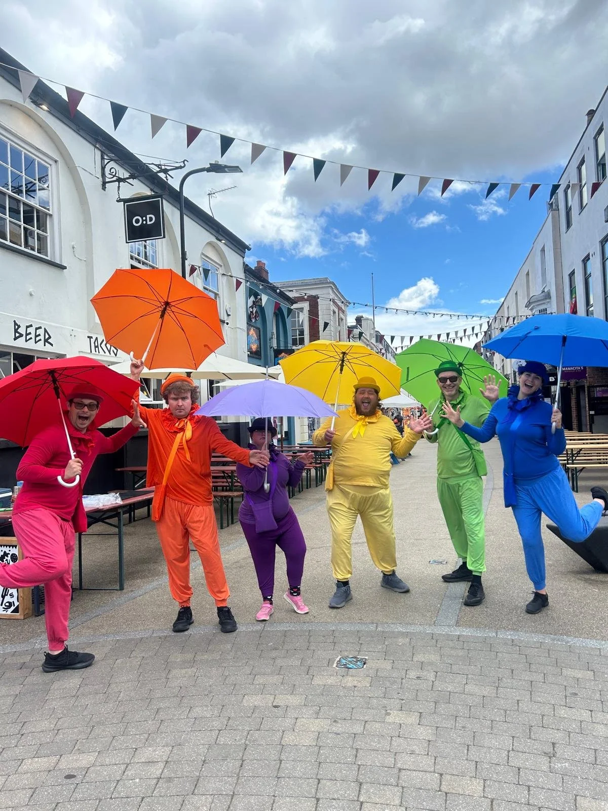 Six performers in rainbow-coloured outfits pose with colourful umbrellas in a pedestrian street decorated with bunting.