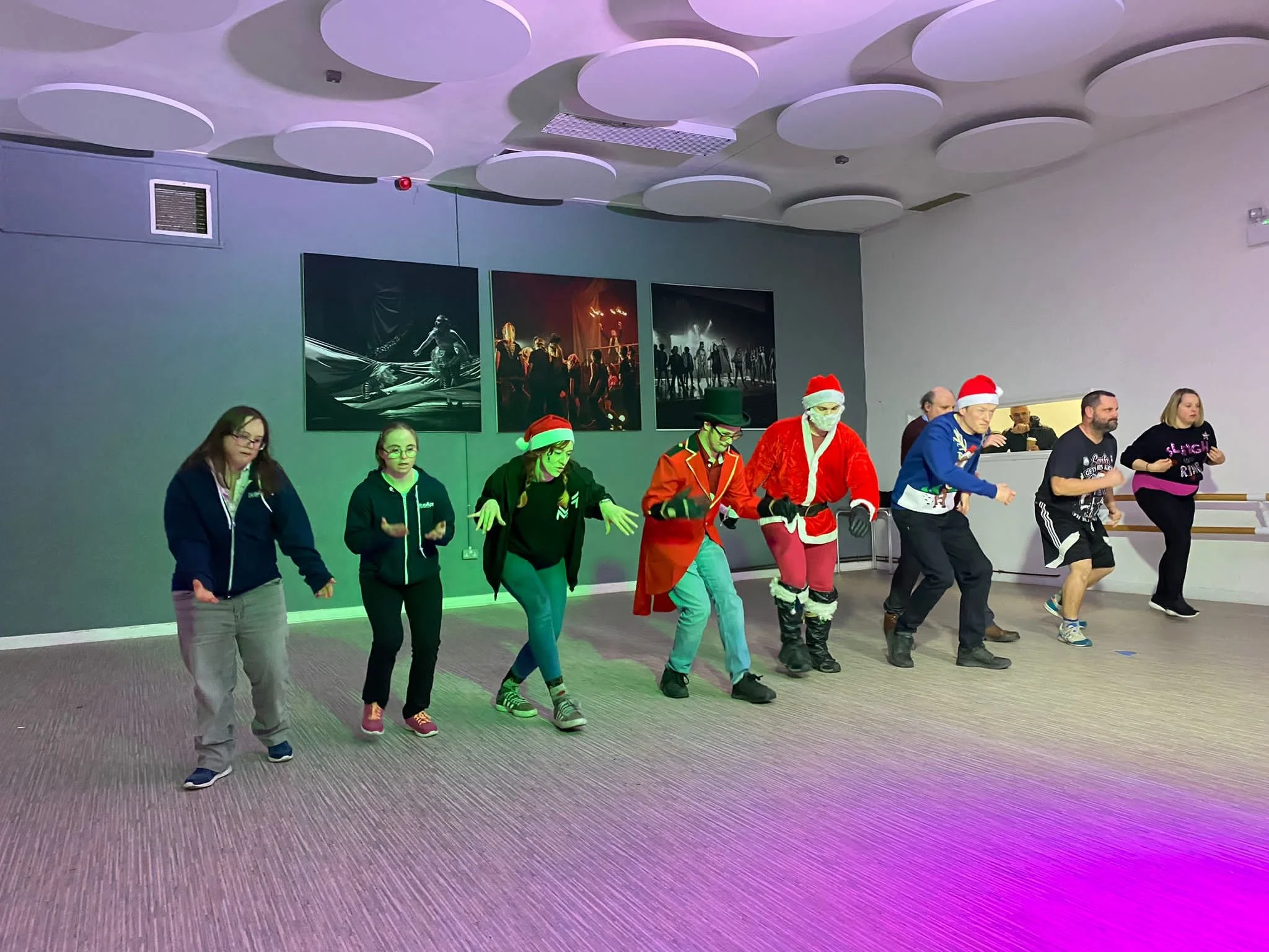 The photo shows dancers in a studio in two photos.  The top one shows them in two rows with the dancers holding their arms outstretched.  The bottom one shows the dances all slightly crouched.  In both photos three of the dancers are wearing Santa ha