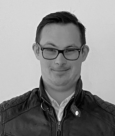 head and shoulders shot of man with short dark hair, wearing glasses and a dark jacket with upturned collar and white shirt showing