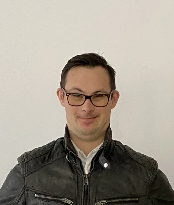 Head and shoulders of a male with dark hair wearing glasses and a dark jacket