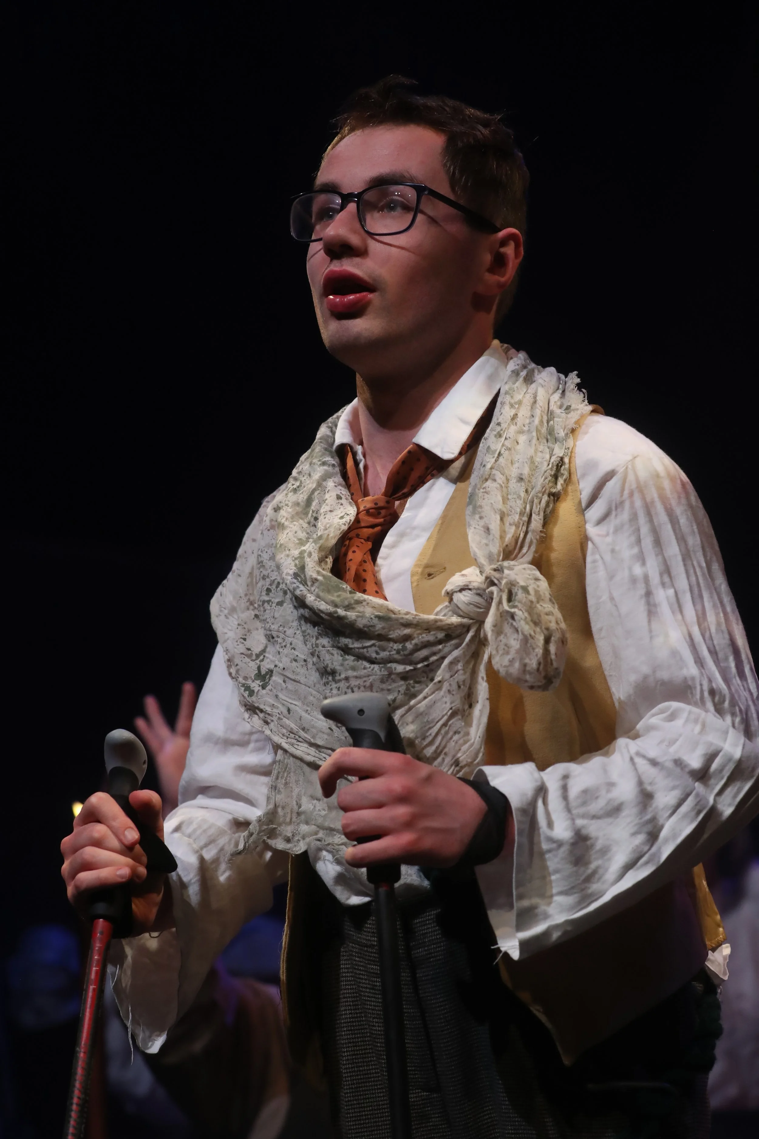 A performer on a dark stage wearing glasses, a white shirt, waistcoat, tie and a loosely draped textured scarf. The performer is holding two mobility aids and is lit by a spotlight, with the background fading into darkness.