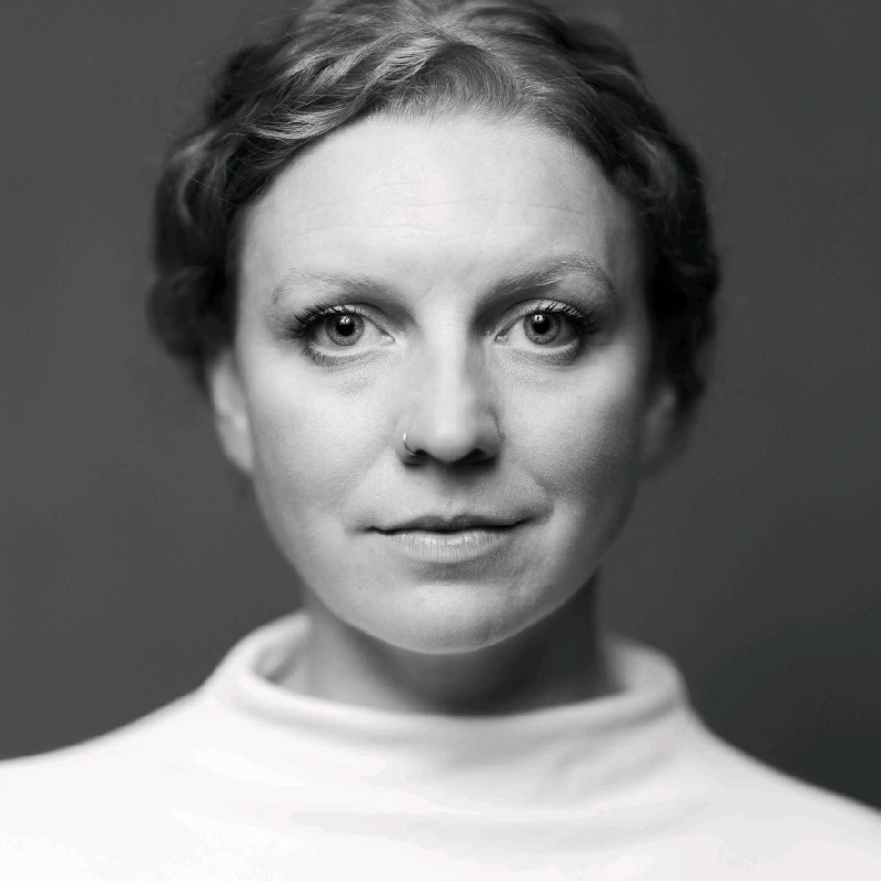A head and shoulder portrait in mono of a woman with her hair braided on top of her head. She wears a white half polo neck top