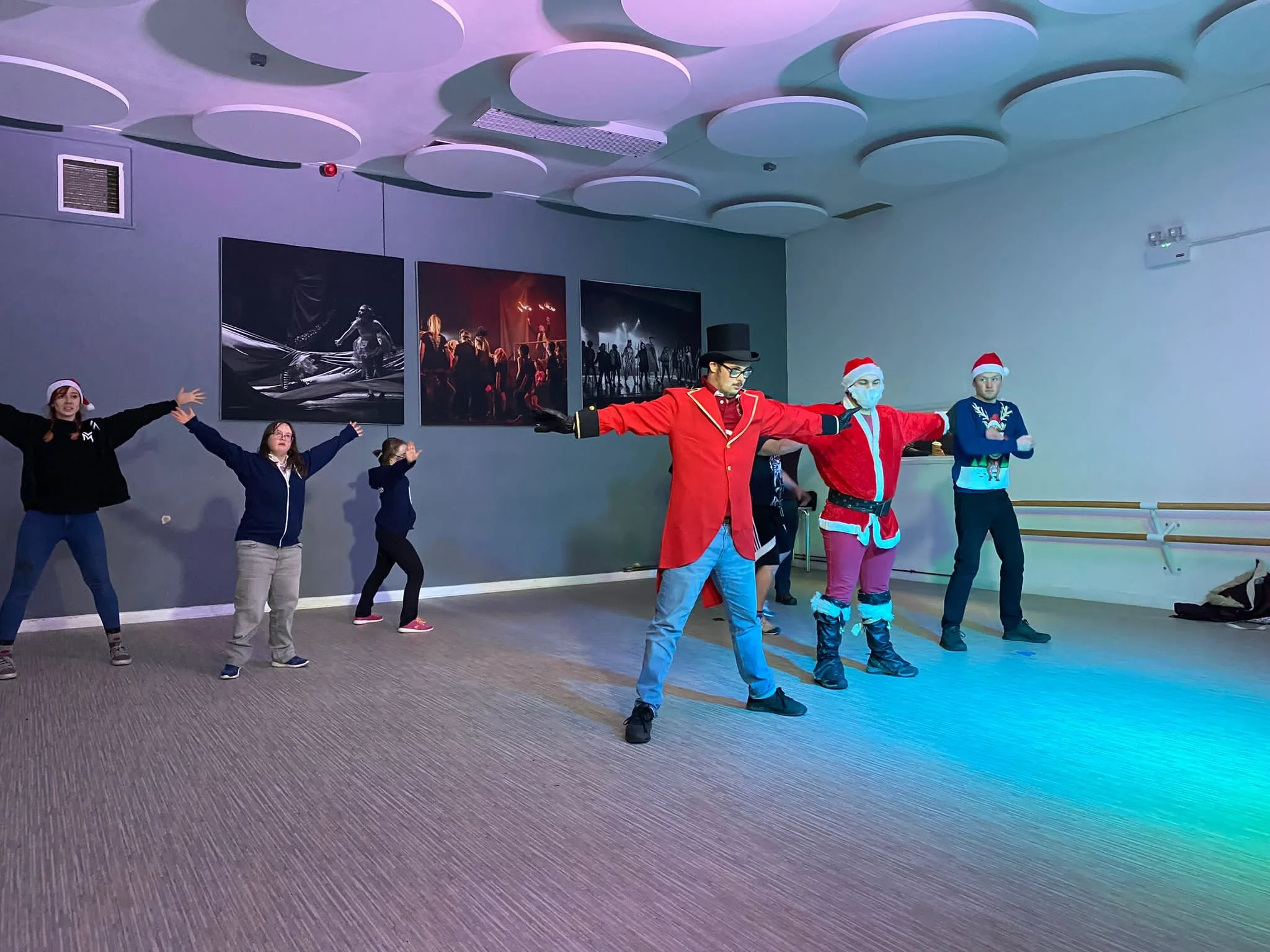 The photo shows dancers in a studio in two photos.  The top one shows them in two rows with the dancers holding their arms outstretched.  The bottom one shows the dances all slightly crouched.  In both photos three of the dancers are wearing Santa ha
