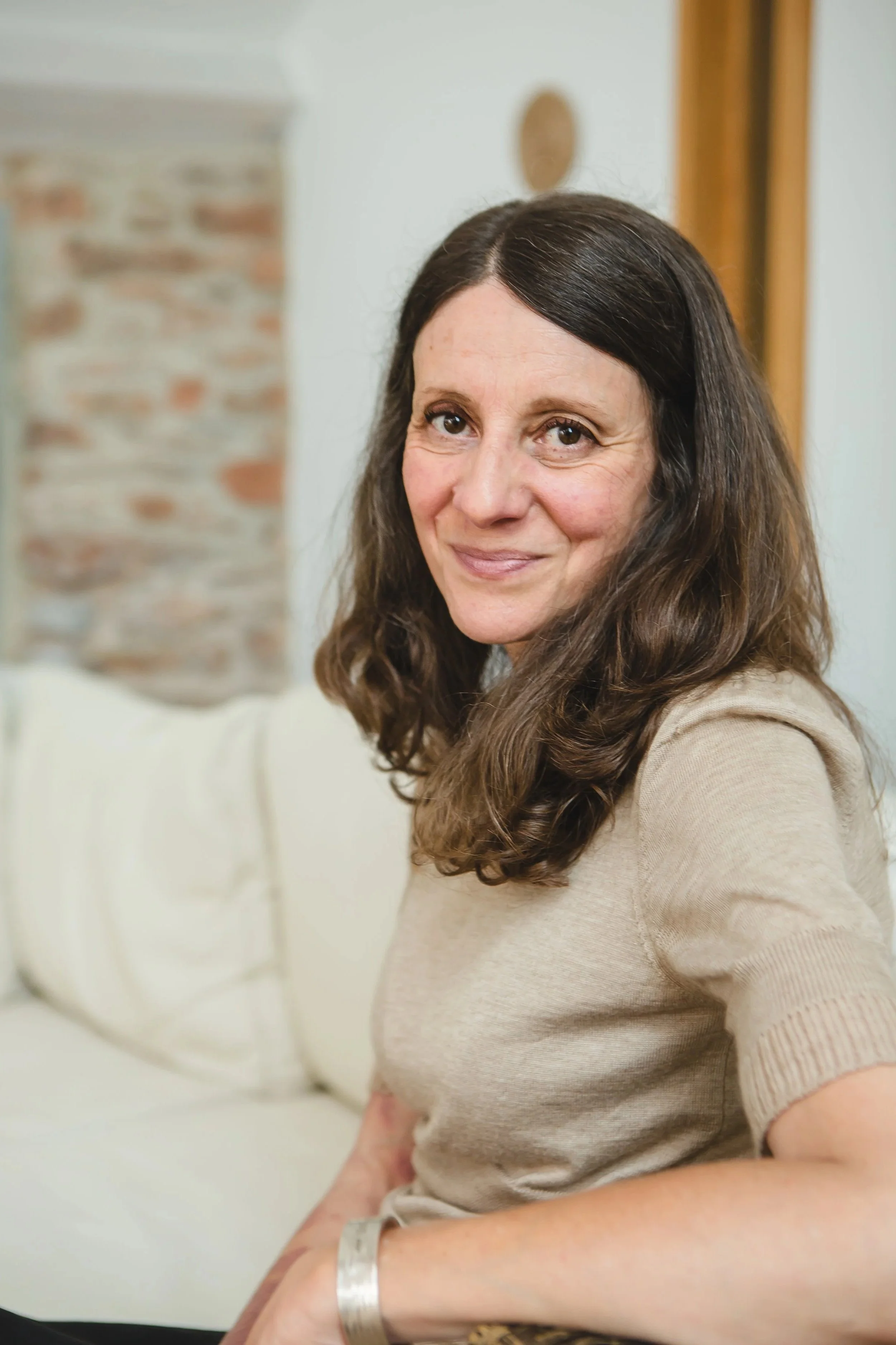 A woman with long brown hair, wearing a beige top, sitting on a cream couch next to a white pillow with a brick pattern. She is smiling and looking slightly to the right of the camera.
