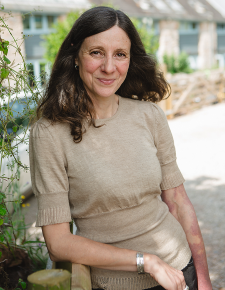A woman with long dark hair sitting outdoors near greenery, wearing a beige sweater and smiling at the camera.