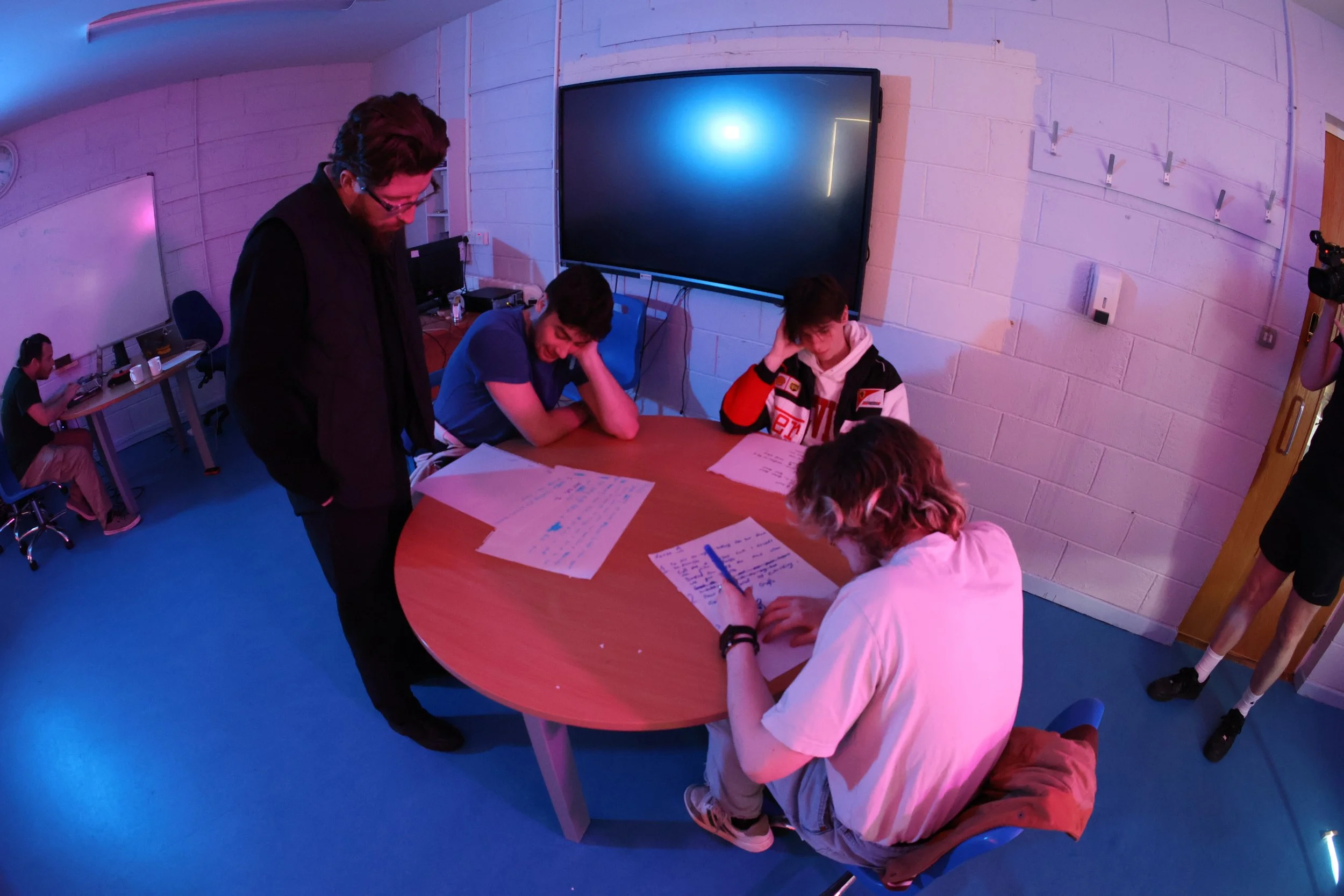 In a classroom with low, purple lighting, Frank McCarthy stands next to a young person at a table and reads their writing. Frank has short, dark hair and a beard, and he wears dark clothes. There are two other young people seated at the table.
