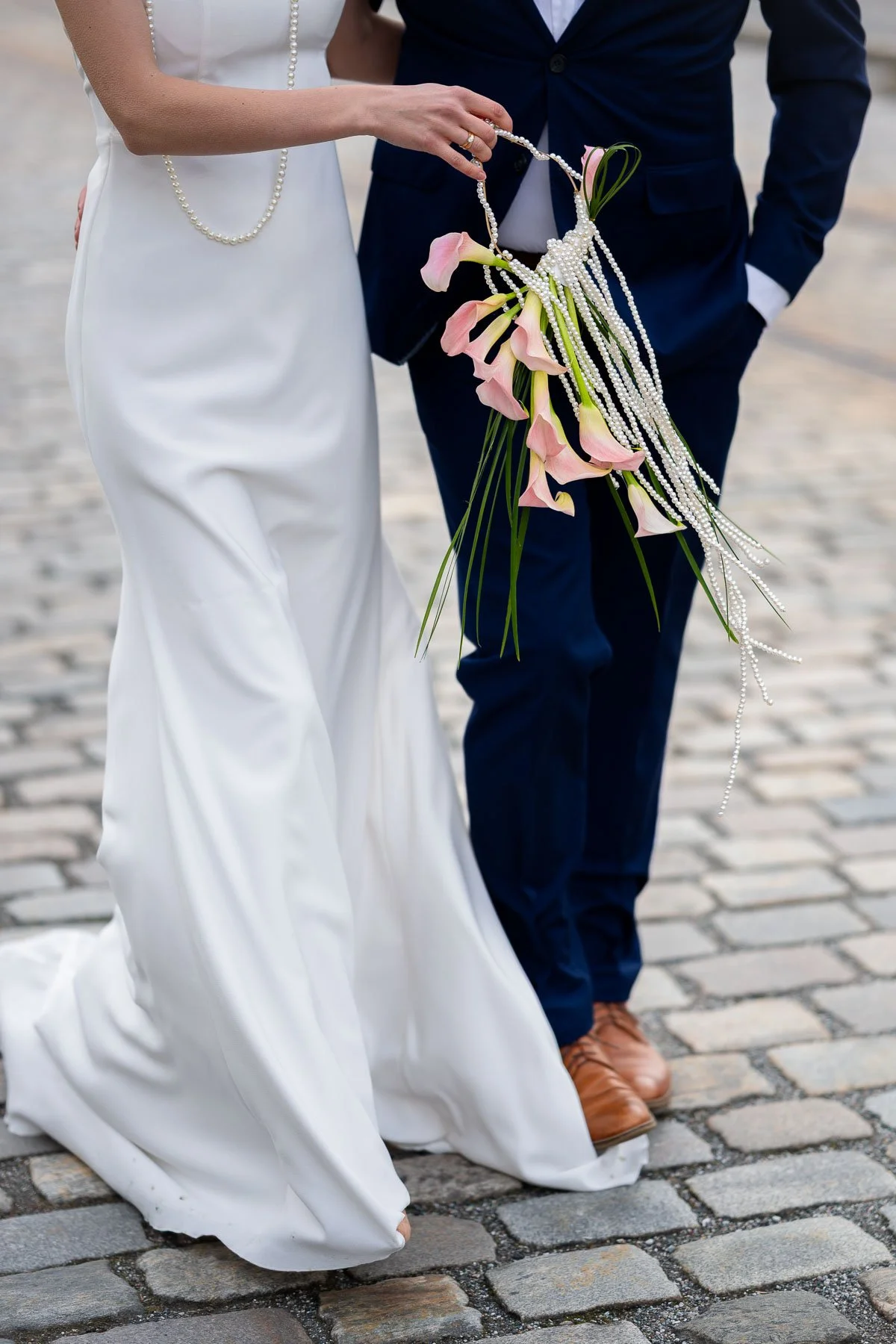 En brud og en brudgom står på brostein, bruden holder en bukett med peoner og perlearmbånd, brudgommen har på seg en mørk blå dress.