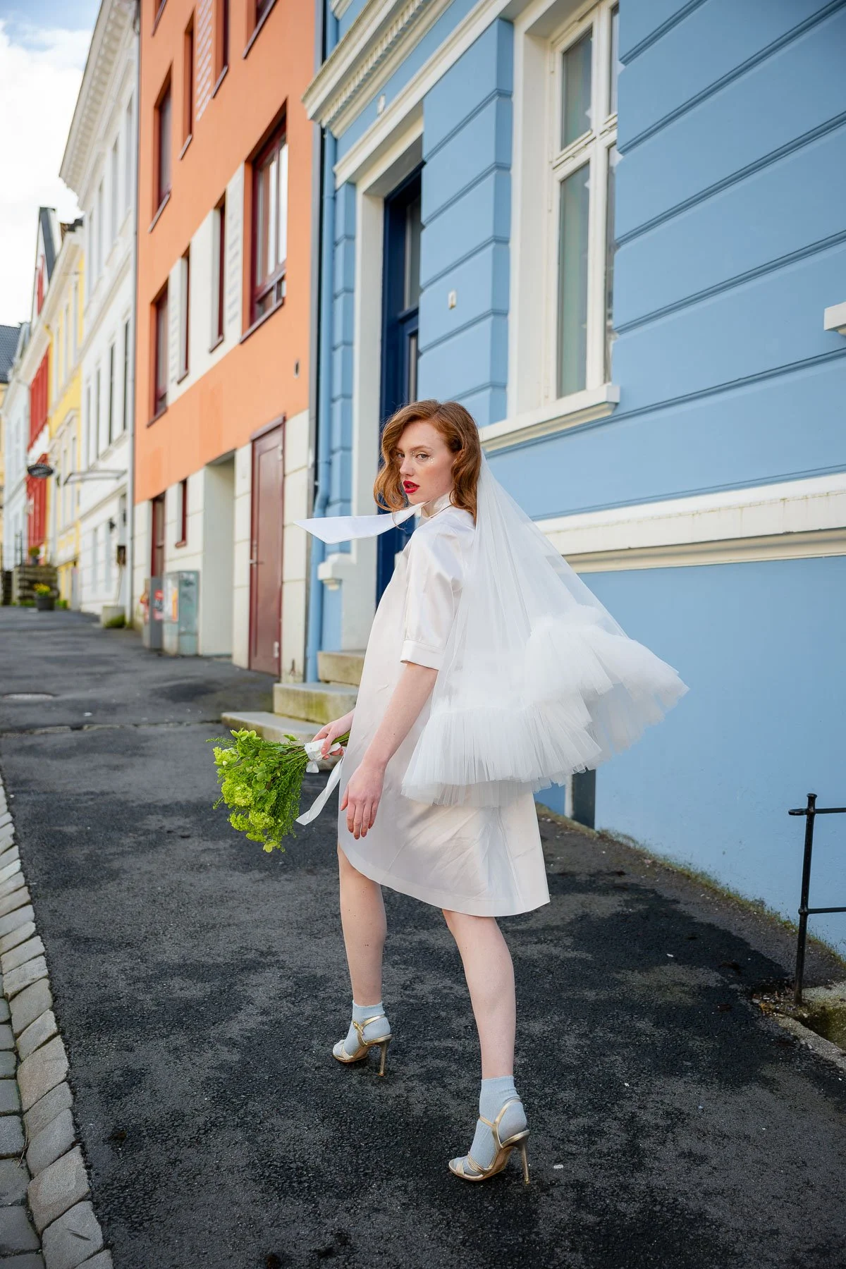 En kvinne i brudeklær står på en gate med fargerike hus. Hun har rødbrunt hår, bærer en hvit kjole og high heels, og holder en hvit flowerbukett. Blikket vender mot kamera.