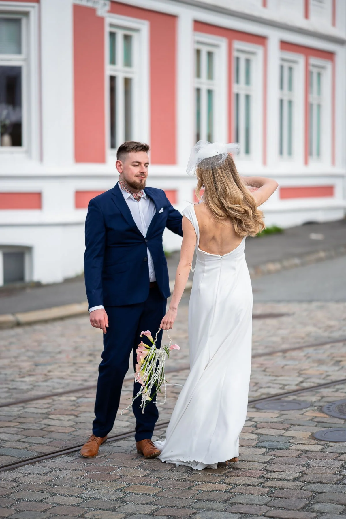 Et brud og en brudgom står på brostein, og ser på hverandre, foran en fargerik bygning.