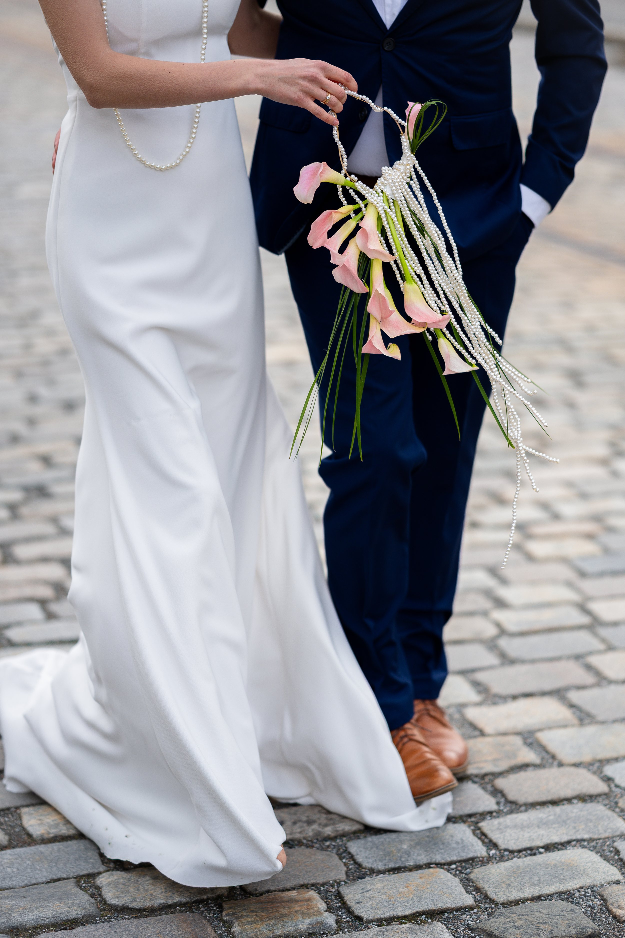 Nærbilde av et bryllup, med en brud i hvit kjole og en brudgom i blå dress, som holder en blomsterbukett og en perleperlekjede sammen, stående på brostein.