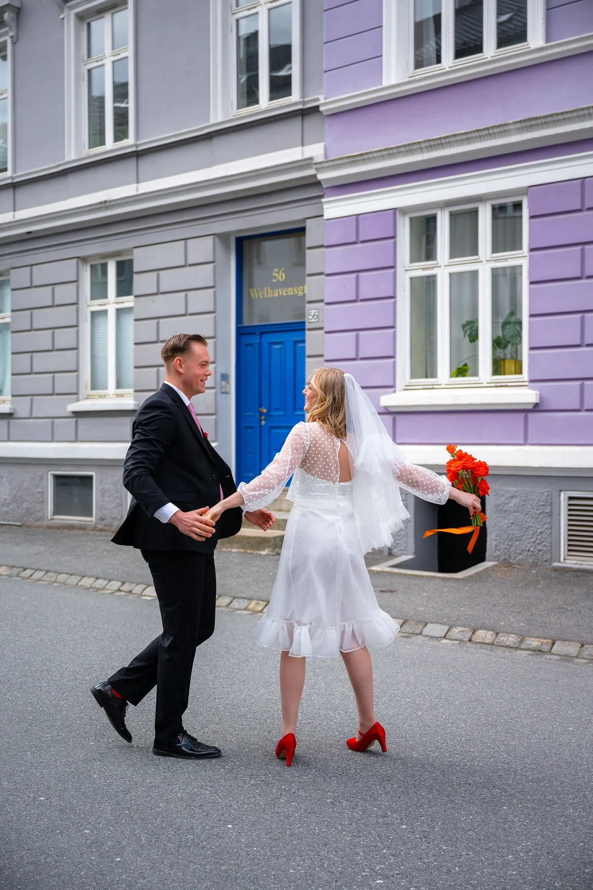 Et brudepar holder hender og danser på gaten foran en fargerik bygning, jenta har på seg en hvit bryllupskjole og røde sko, mens fyren har en svart dress og rosa slips.