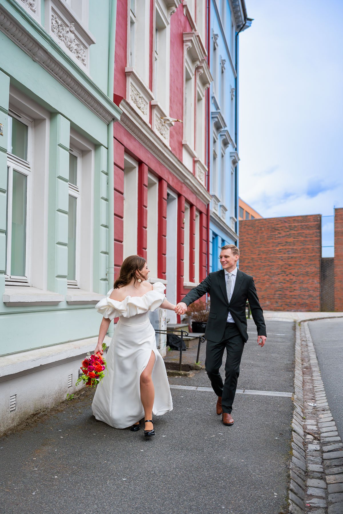 Et par kledd i bryllupsantrekk går hånd i hånd langs en gate med fargerike bygninger. Damen har på seg en hvit brudekjole med åpen skulder og høy splitt, og holder en blomsterbukett. Herren er i en svart dress.