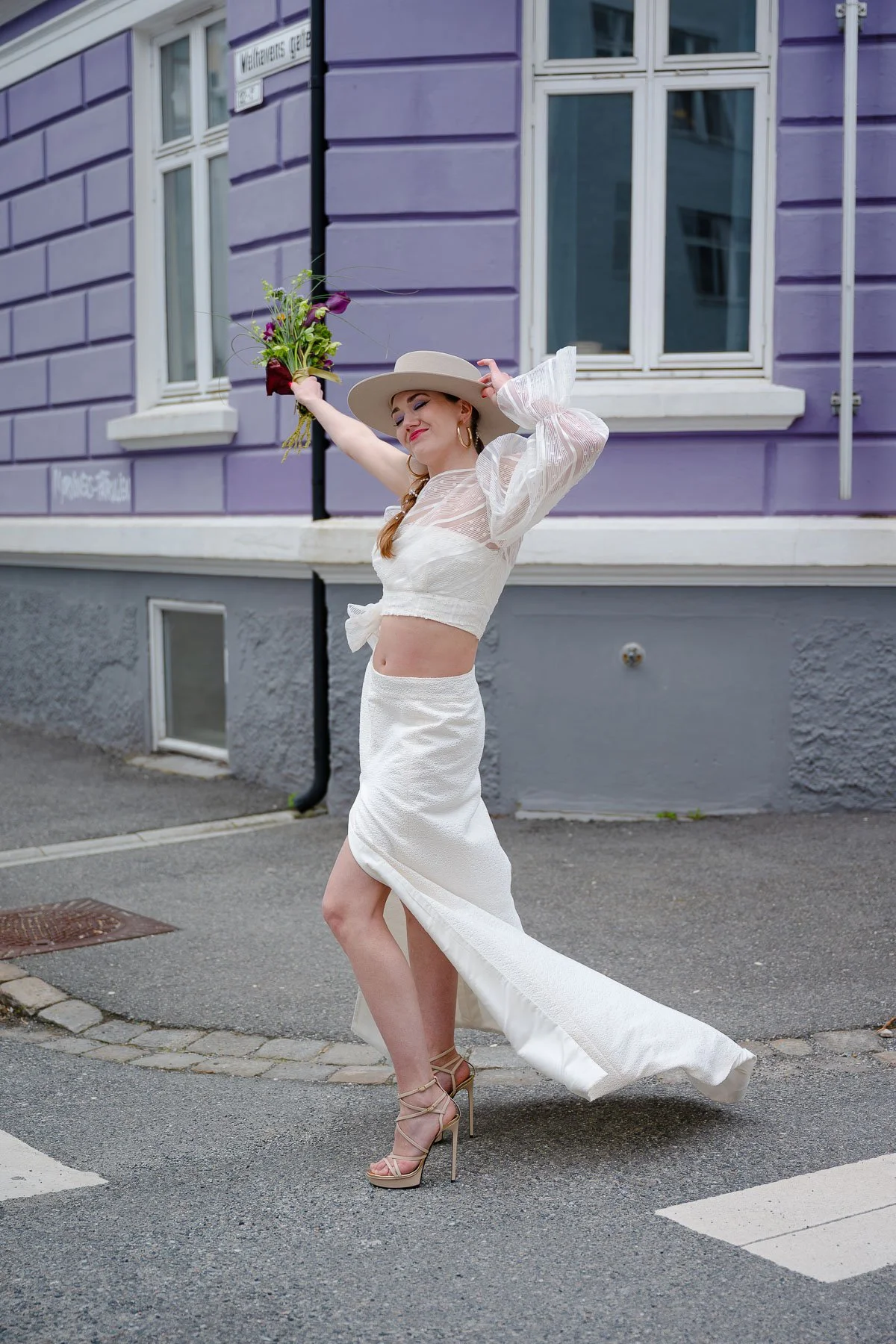 Kvinne i hvit kjole og hat holder blomster på gaten, smilende.