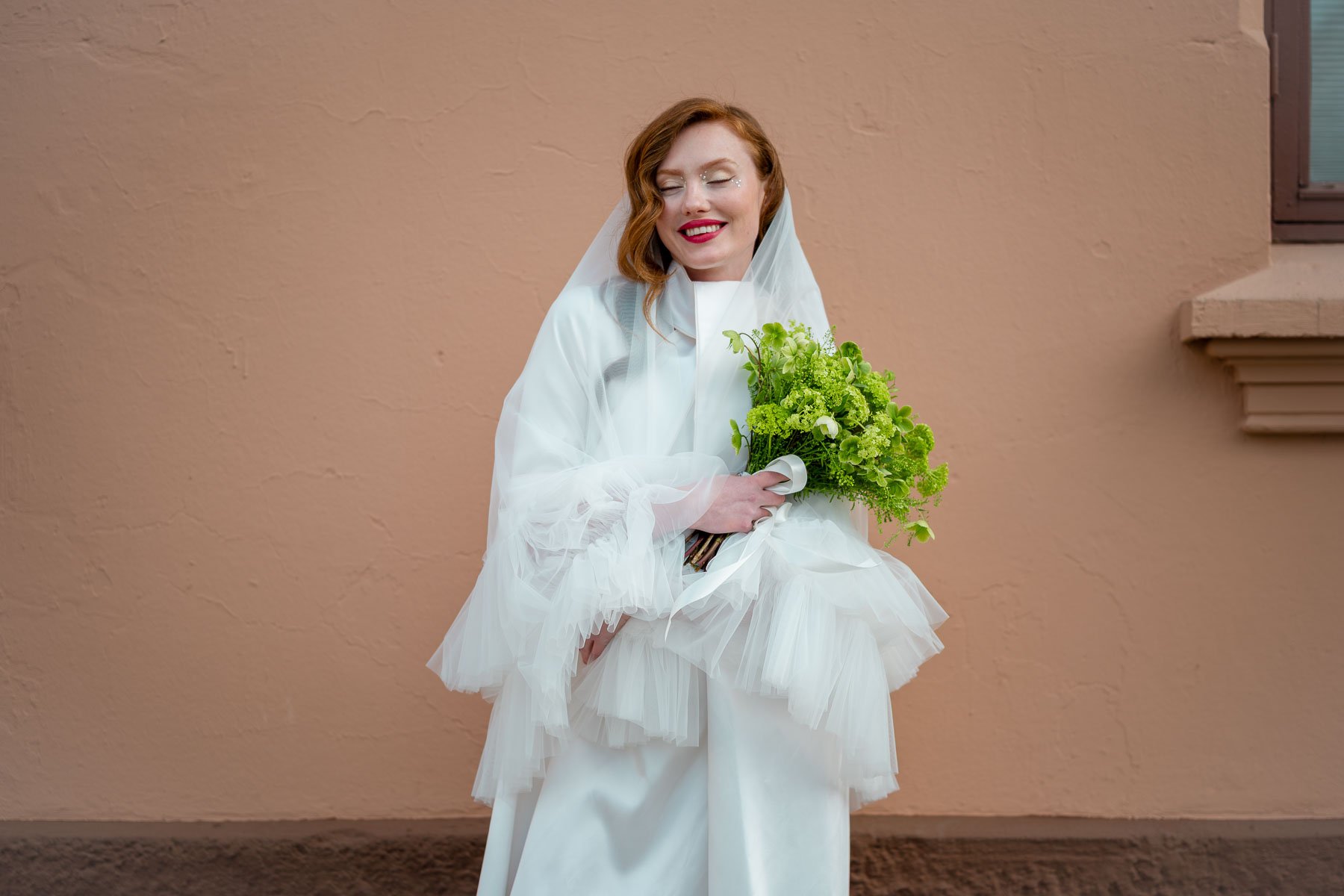 En kvinne i brudekjole holder en grønn bukett, smiler med lukkede øyne og står mot en beige vegg.