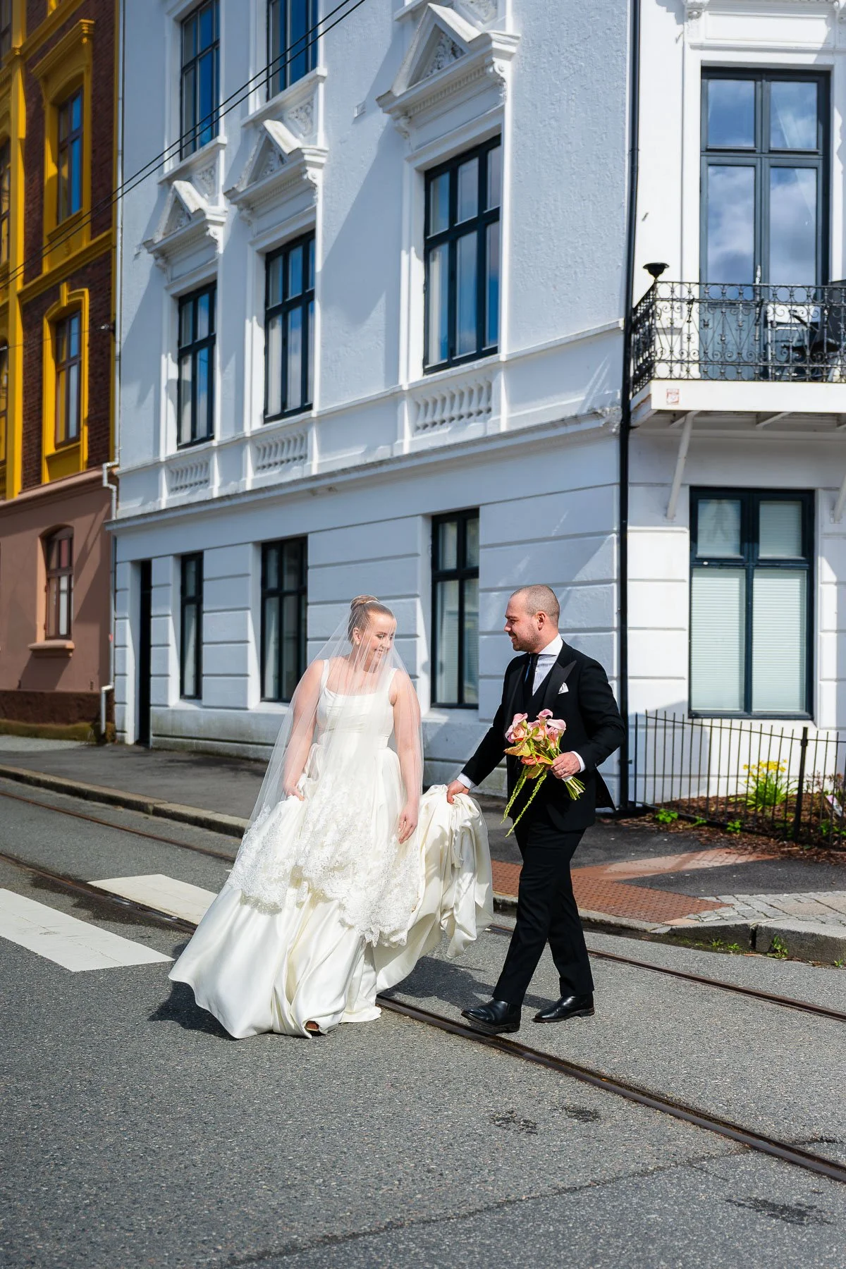 En brud og brudgom går over en gate, der brudgommen holder en bukett blomster. Bruden har på seg en hvit brudekjole og brudekveil. De smiler mot hverandre foran hvite og fargede bygninger.