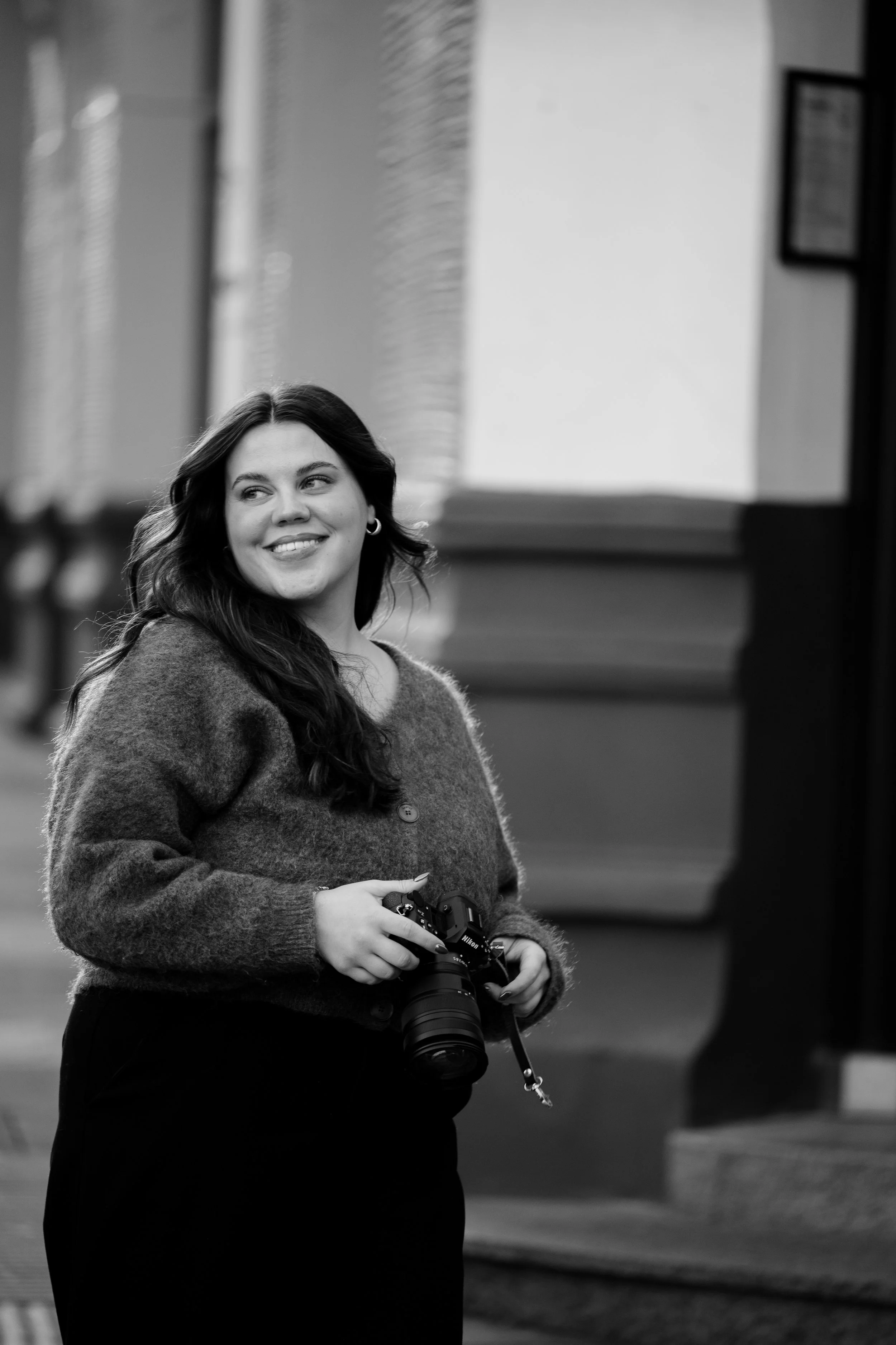 En ung kvinne med langt hår smiler og holder et kamera, står innendørs.