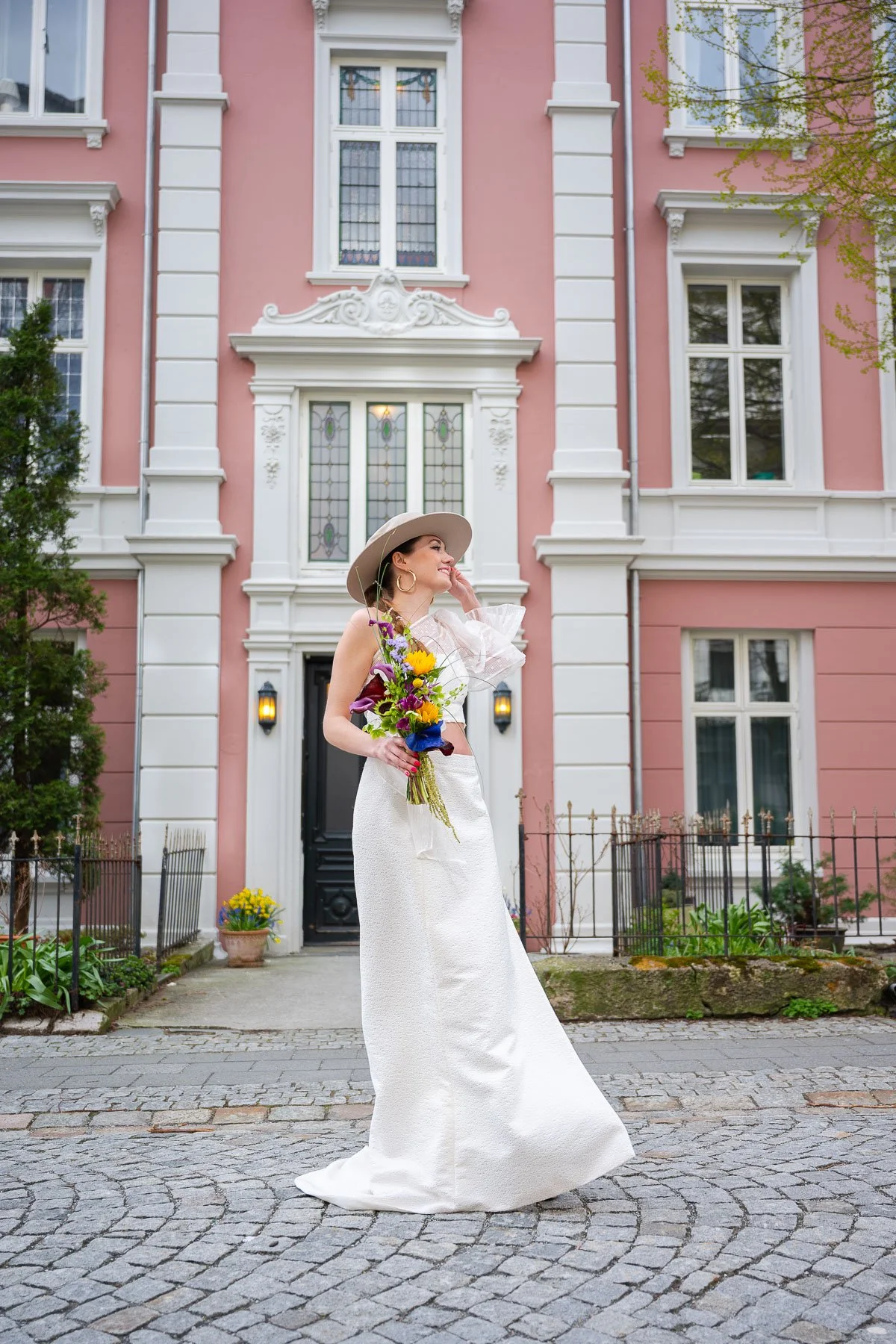 En kvinne i en brudekjole holder en fargerik blomsterbukett foran en rosa bygning med store vinduer.