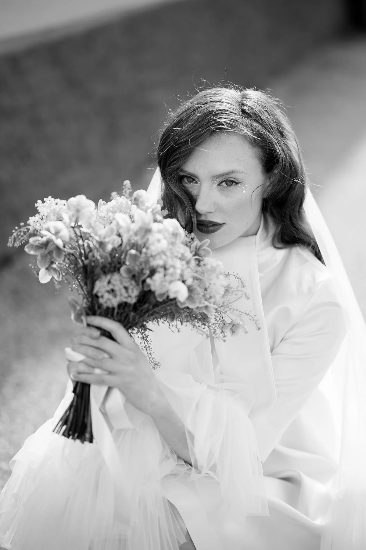 Svart-hvitt bilde av en kvinne med langt hår som holder en blomsterbukette, ser direkte inn i kameraet, med smykker ved øynene og mørk leppestift, i en utendørs setting.