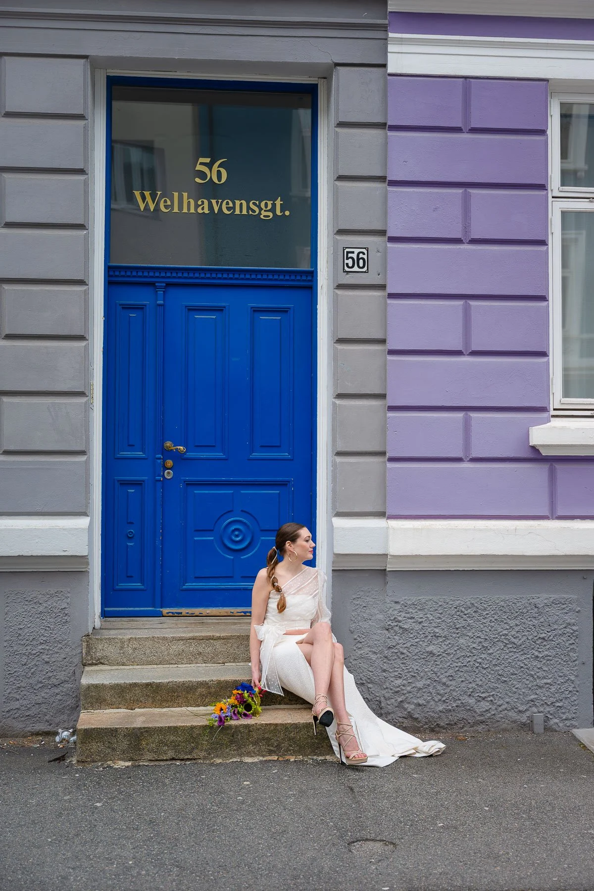 En kvinne i en hvit kjole og høye hæler sitter på trappetrinn foran en blå dør med nummer 56. Ved siden av henne ligger en blomsterbukett. En bygning med grå og lilla vegger er synlig i bakgrunnen.
