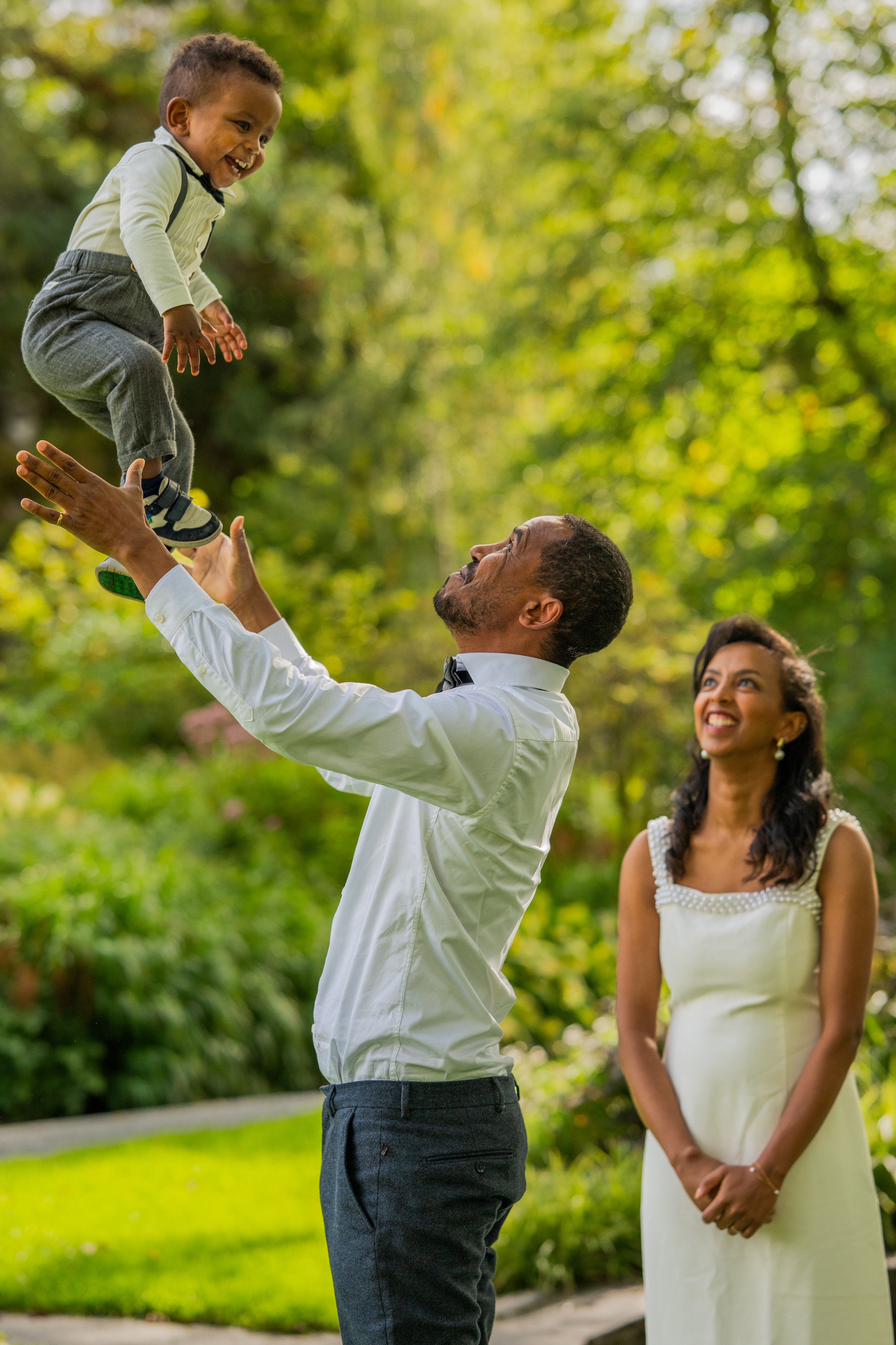 En mann kaster en liten gutt opp i luften mens en kvinne står ved siden av og smiler i en hage med grønne trær.