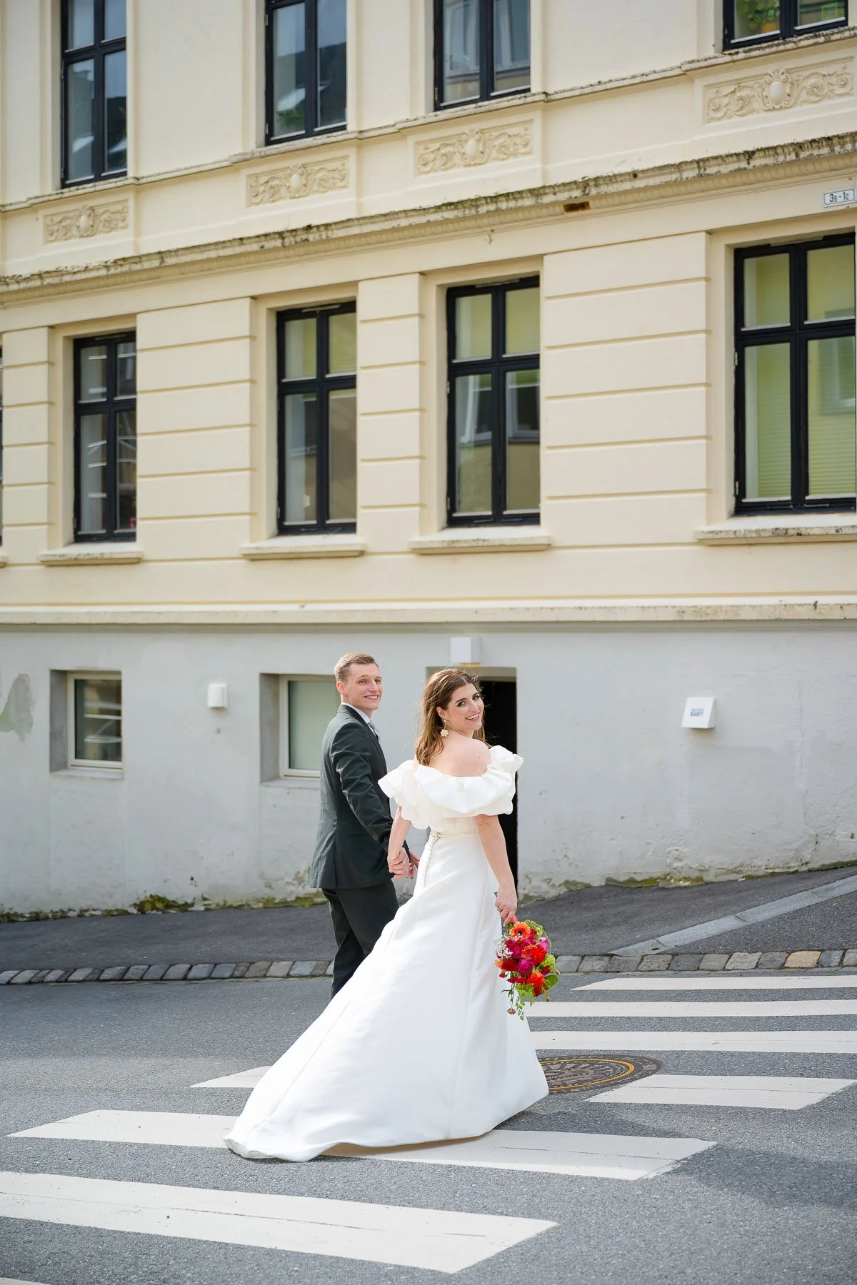 Et brudepar går hånd i hånd over et fotgjengerovergang på gaten, med en eldre bygning i bakgrunnen.