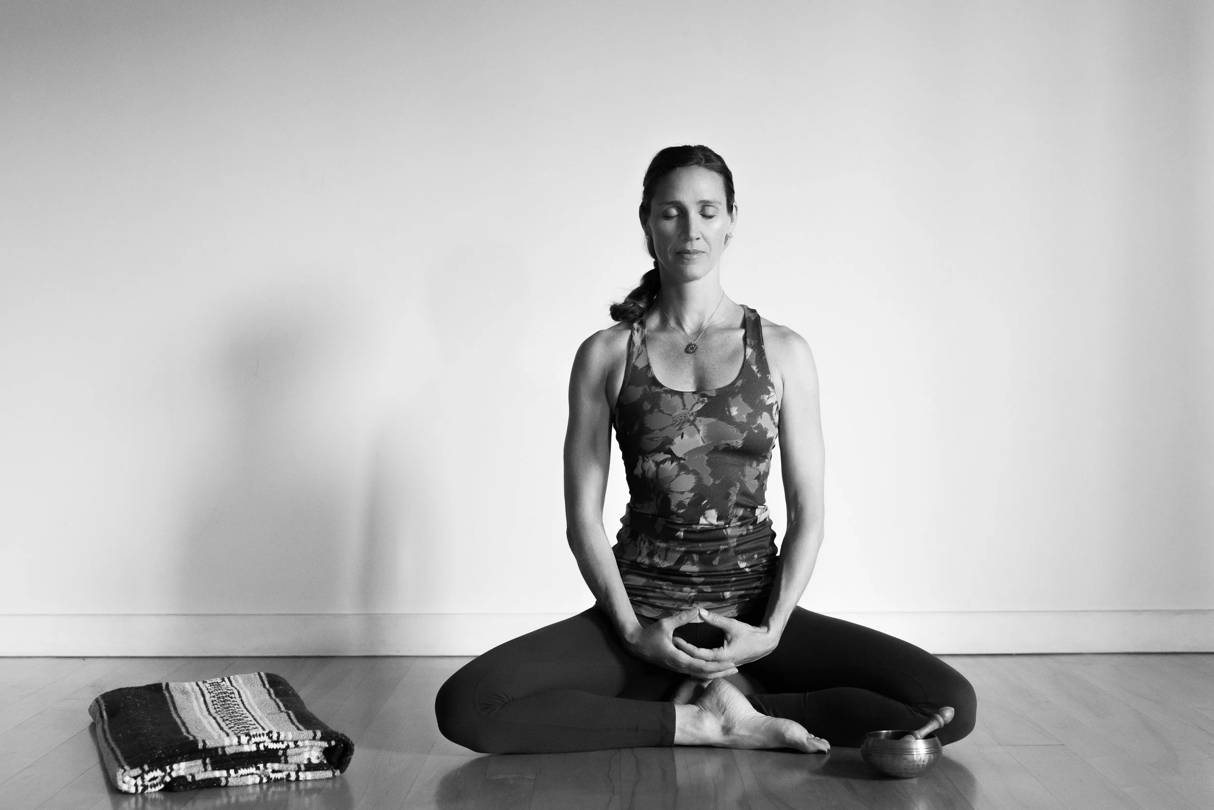 Vivica Schwartz practicing yoga indoors, sitting cross-legged on a yoga mat, with her hands in a prayer position on her lap, eyes closed, and a serene expression.