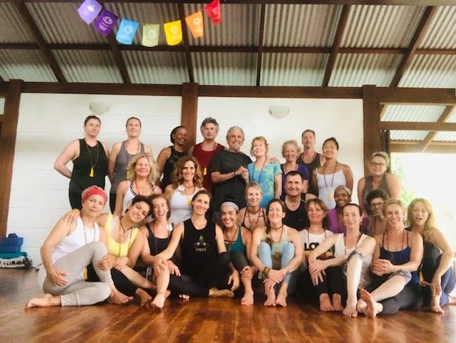 Group of people in a yoga studio with colorful flags hanging from the ceiling, posing for a group photo after a class.