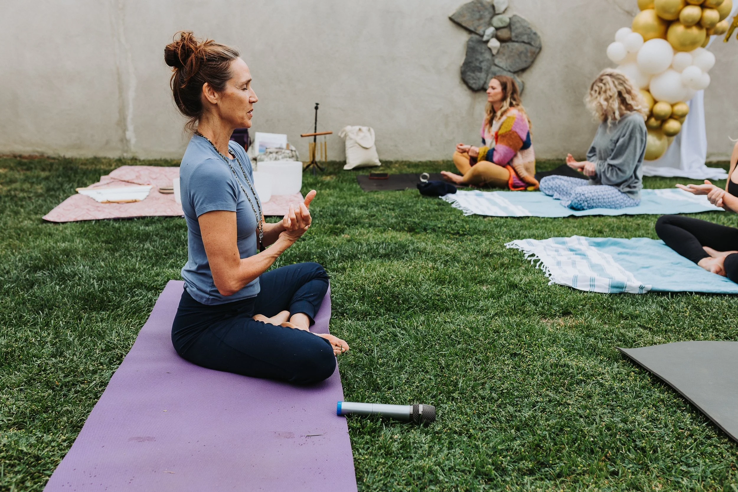 Vivica Schwartz participating in an outdoor yoga or meditation session on mats, with a woman leading the practice, and balloons in the background.
