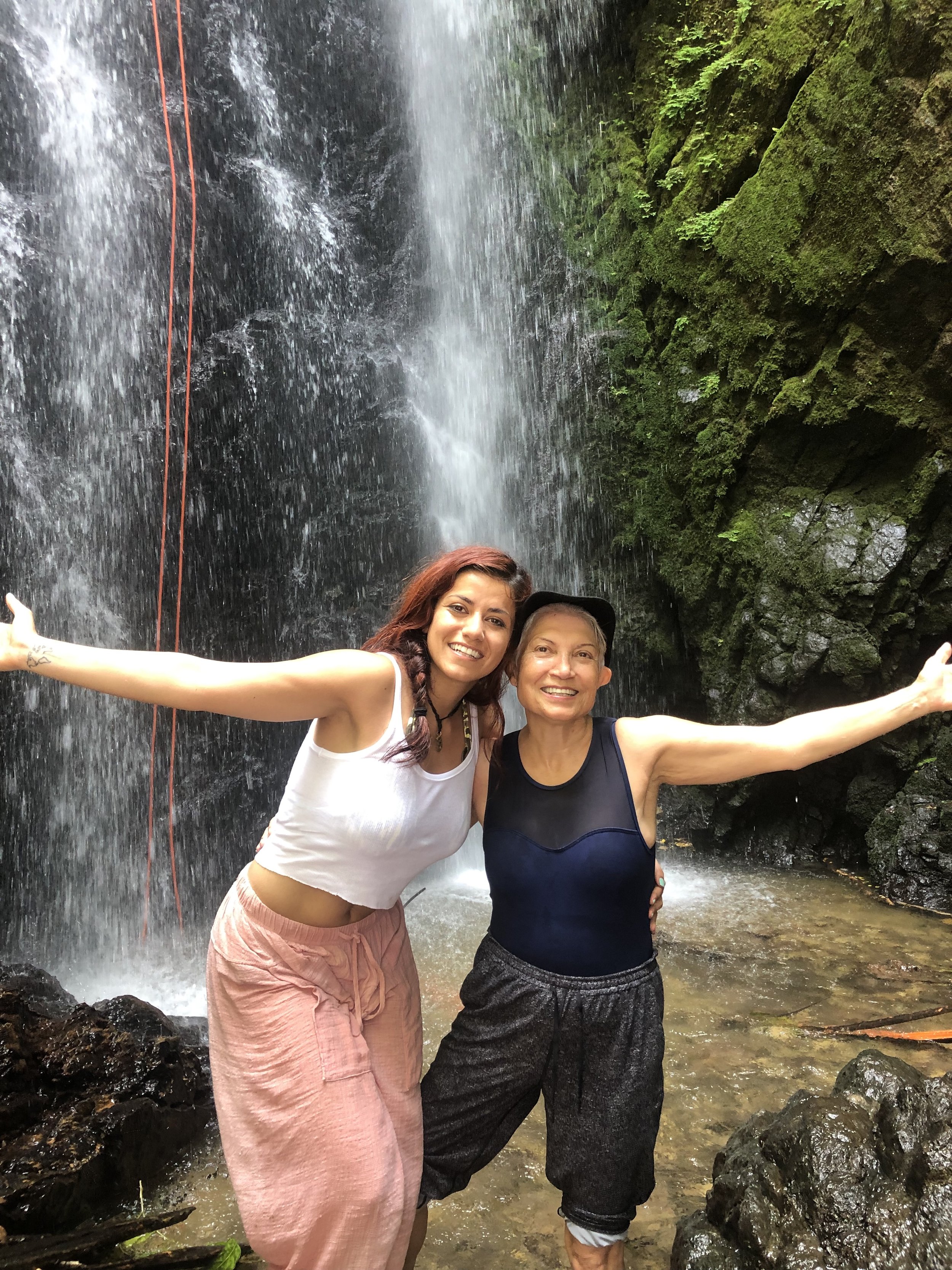 Two women smiling and extending their arms in front of a waterfall with moss-covered rocks and greenery. Yoga retreat in Costa Rica led by Vivica Schwartz. 